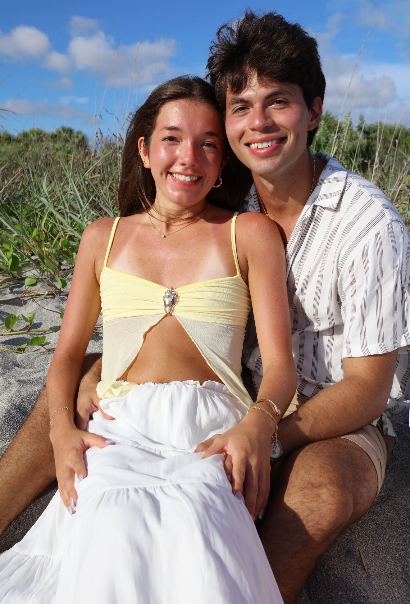 A smiling young couple poses together on a sandy beach, the woman in a yellow crop top and white skirt.