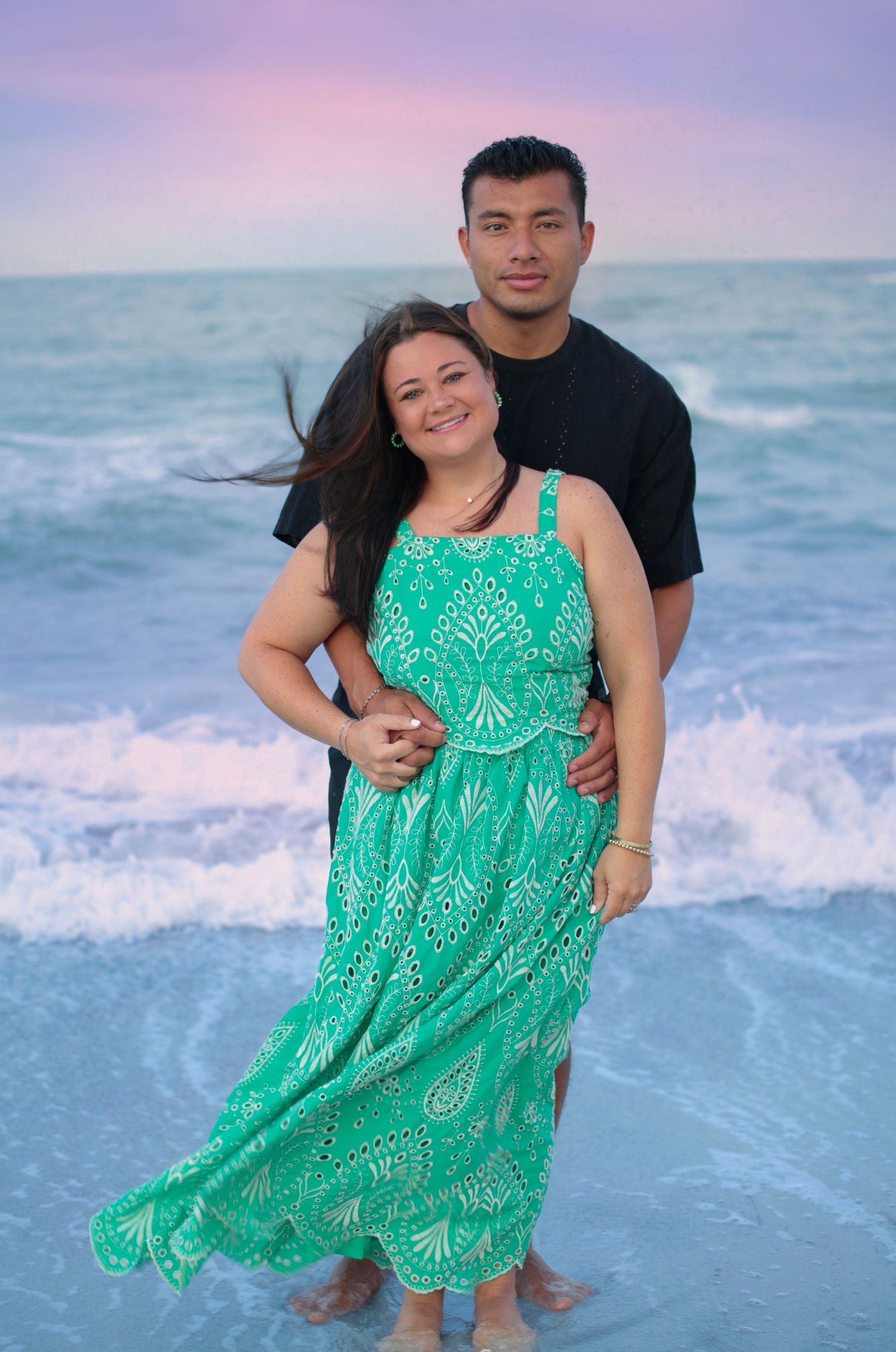 Couple embracing on a beach at sunset, woman in green maxi dress, man in black shirt, ocean waves behind them.