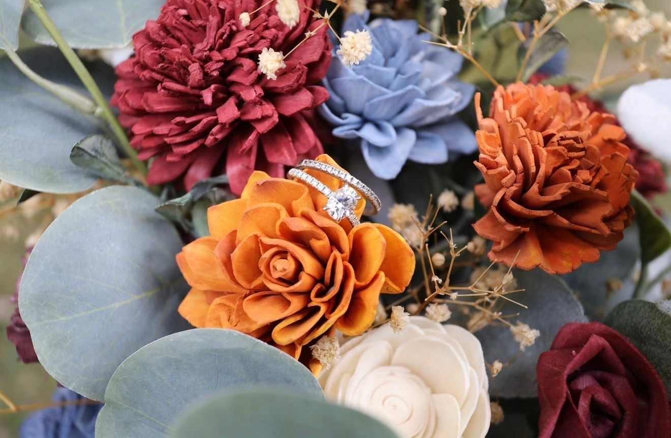 Elegant sola wood flowers in burgundy, blue, rust, and cream with a diamond ring nestled among eucalyptus leaves.