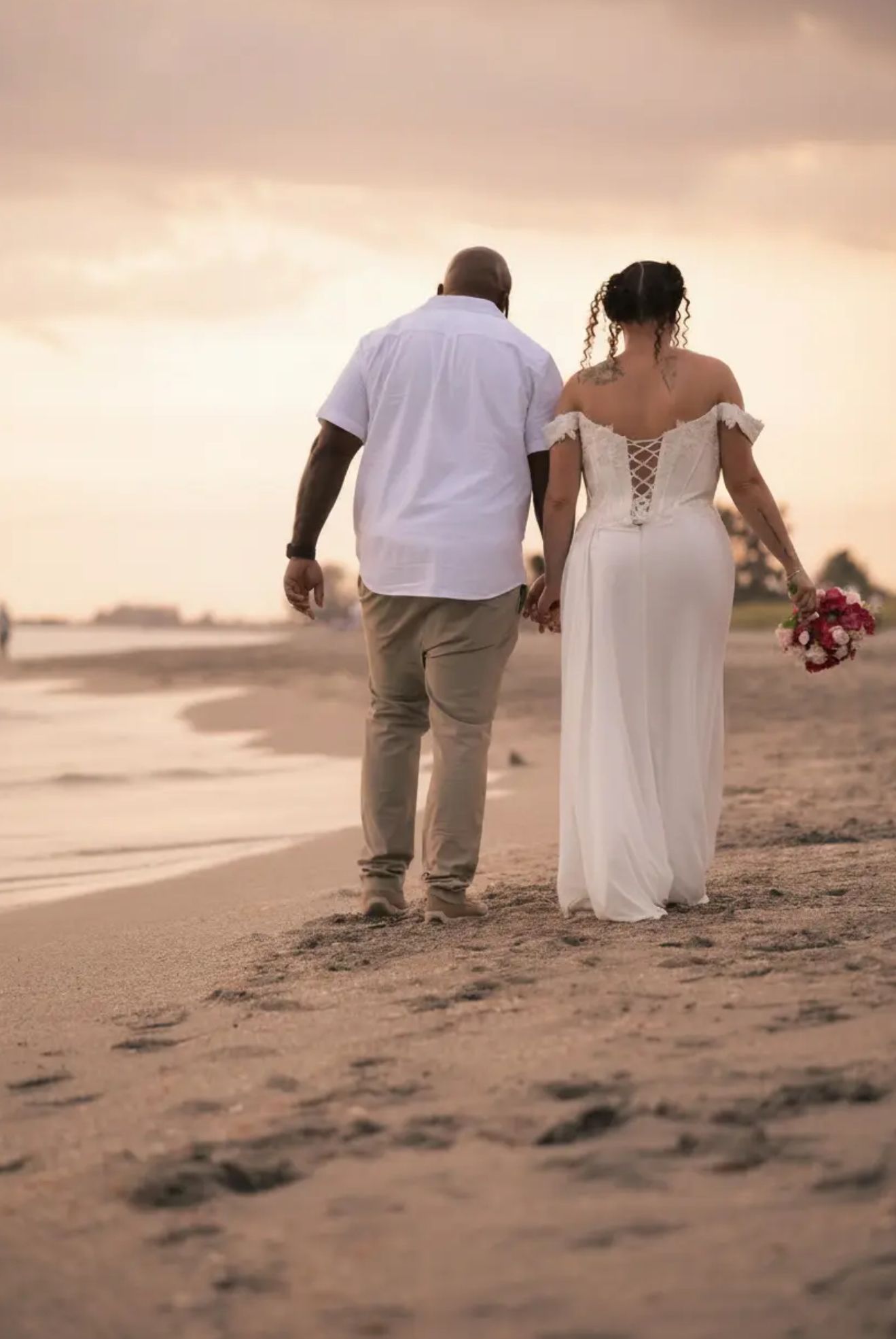 Newlywed couple holding hands walking on a sandy beach at sunset, bride in white off-shoulder gown holding bouquet.