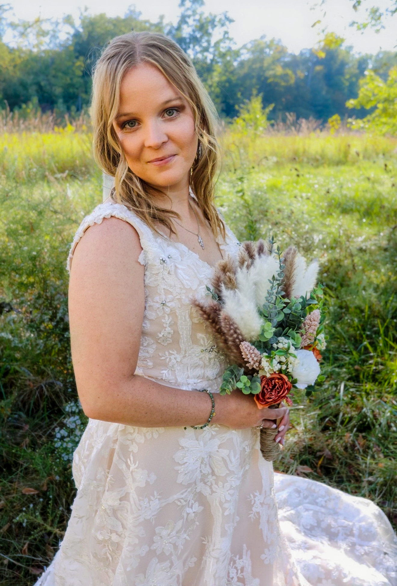 Blonde bride in lace wedding gown holds boho bouquet with dried pampas grass and red roses in a sunlit meadow.