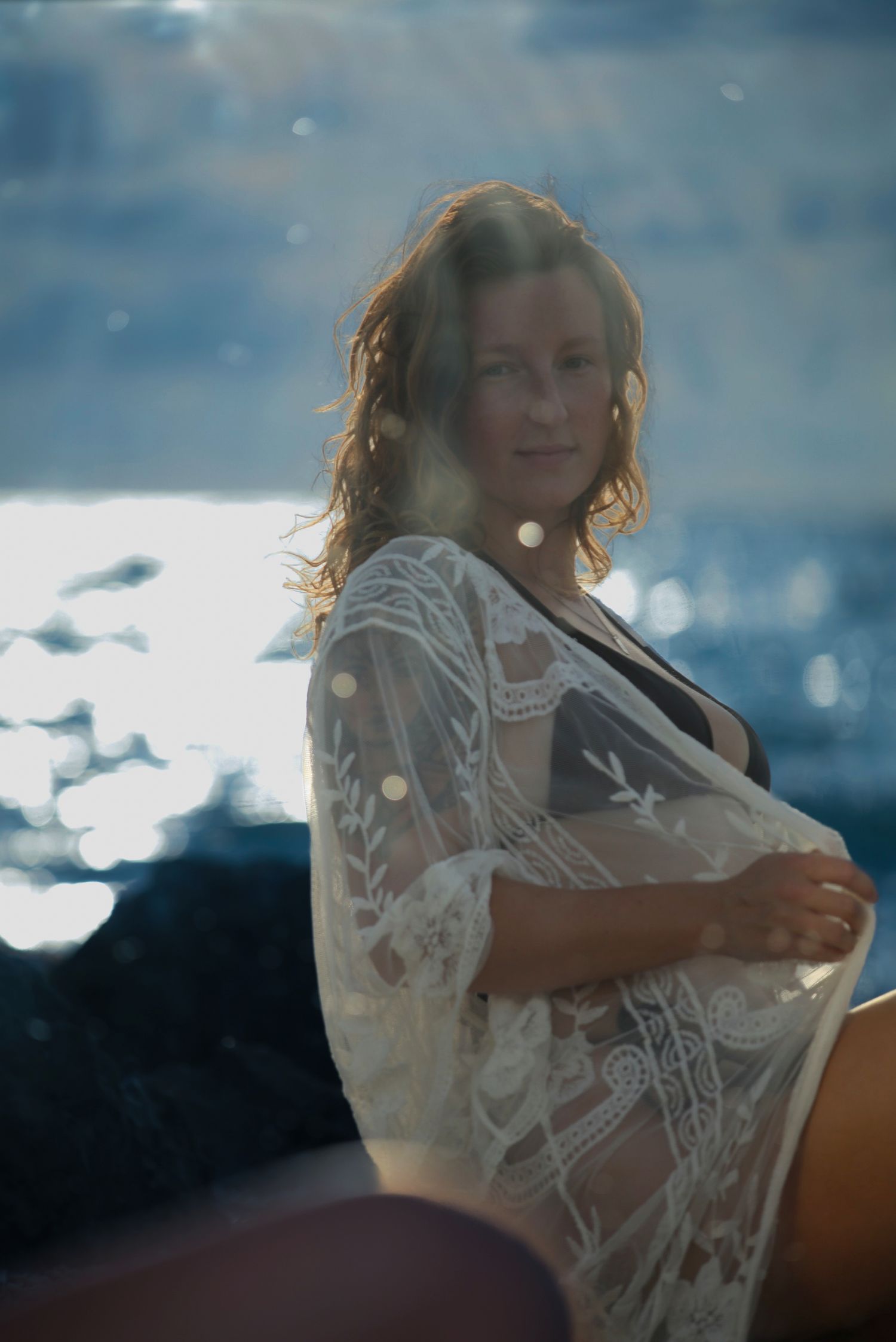 Woman in white lace cover-up poses by the ocean with sunlight reflecting off the water behind her.