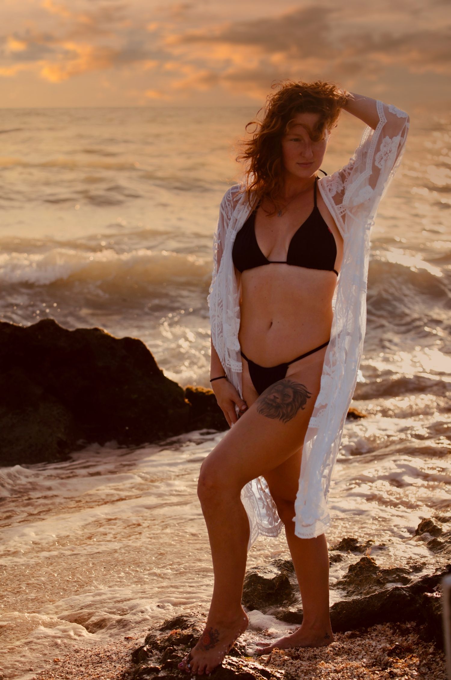 Woman in black bikini and sheer white cover-up stands on rocks with ocean waves at golden sunset.