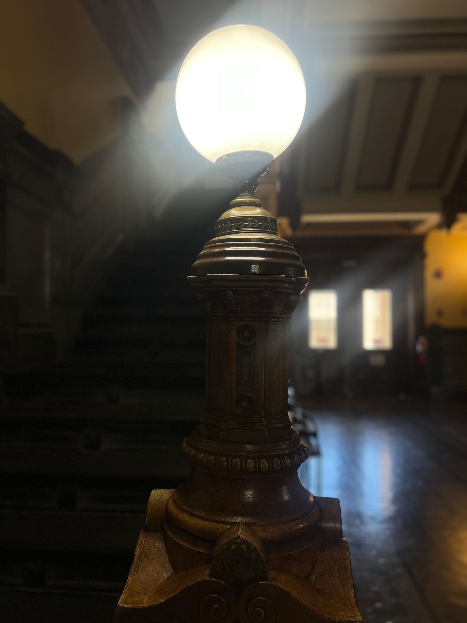 Ornate vintage brass lamp with glowing globe on decorative base inside historic building interior.