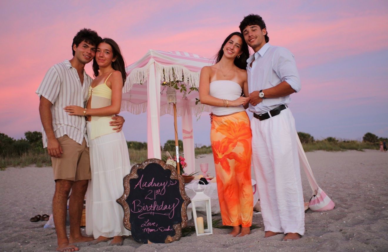 Two couples pose on a beach at sunset near a decorated canopy and birthday chalkboard sign.