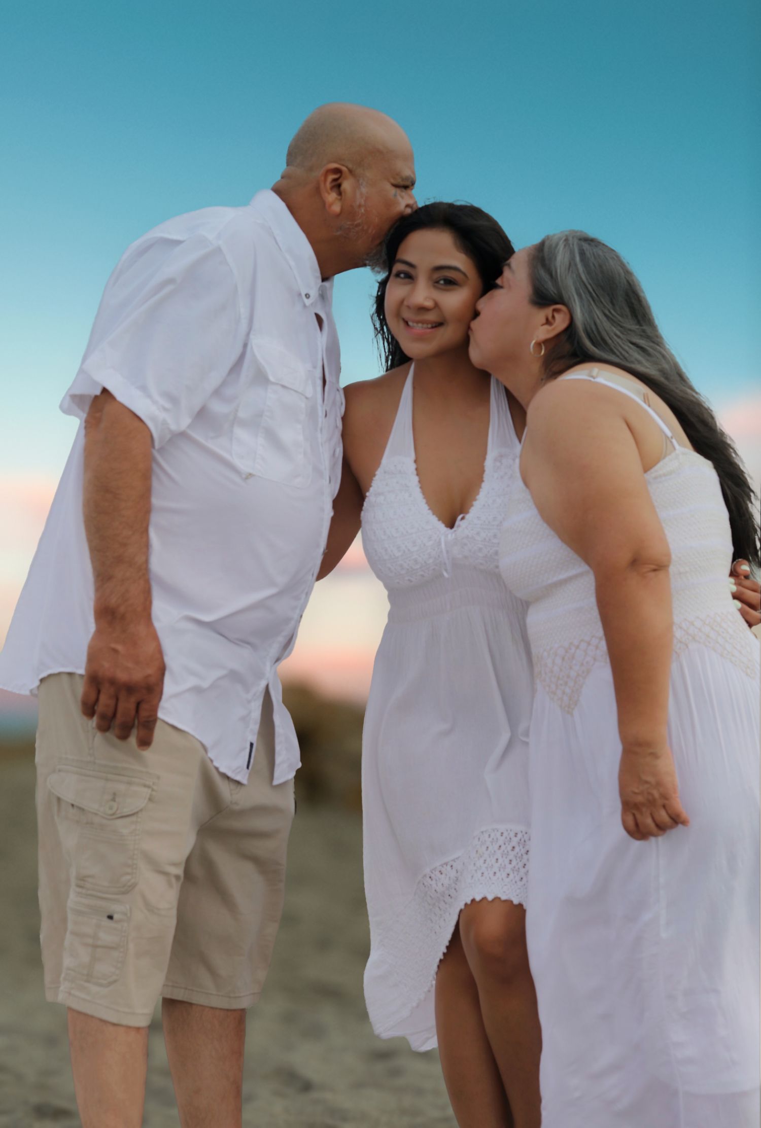 Elderly couple kisses their smiling daughter on the cheeks on a beach at sunset.