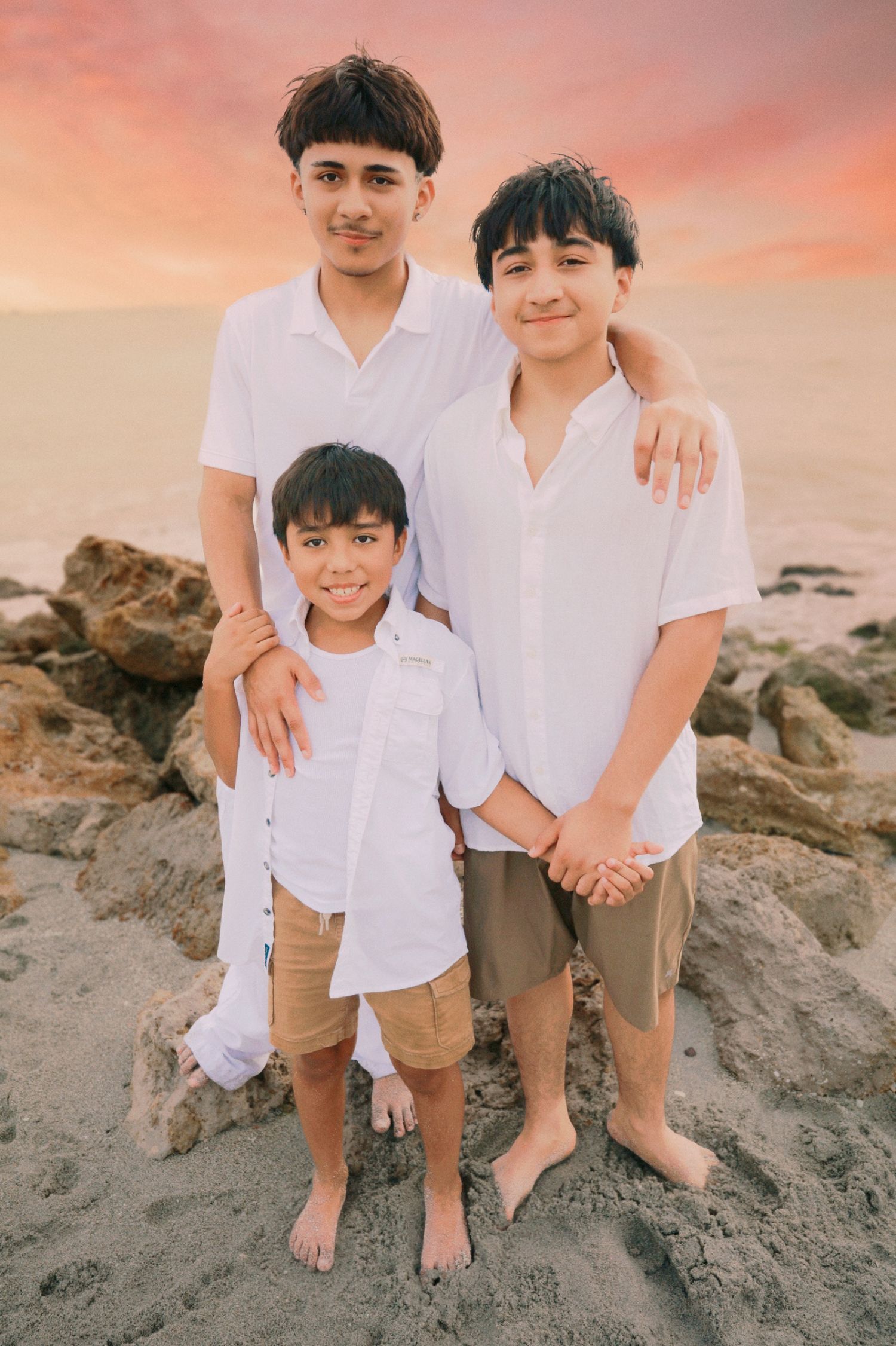 Three boys in white shirts and tan shorts pose together on a sandy beach at sunset.