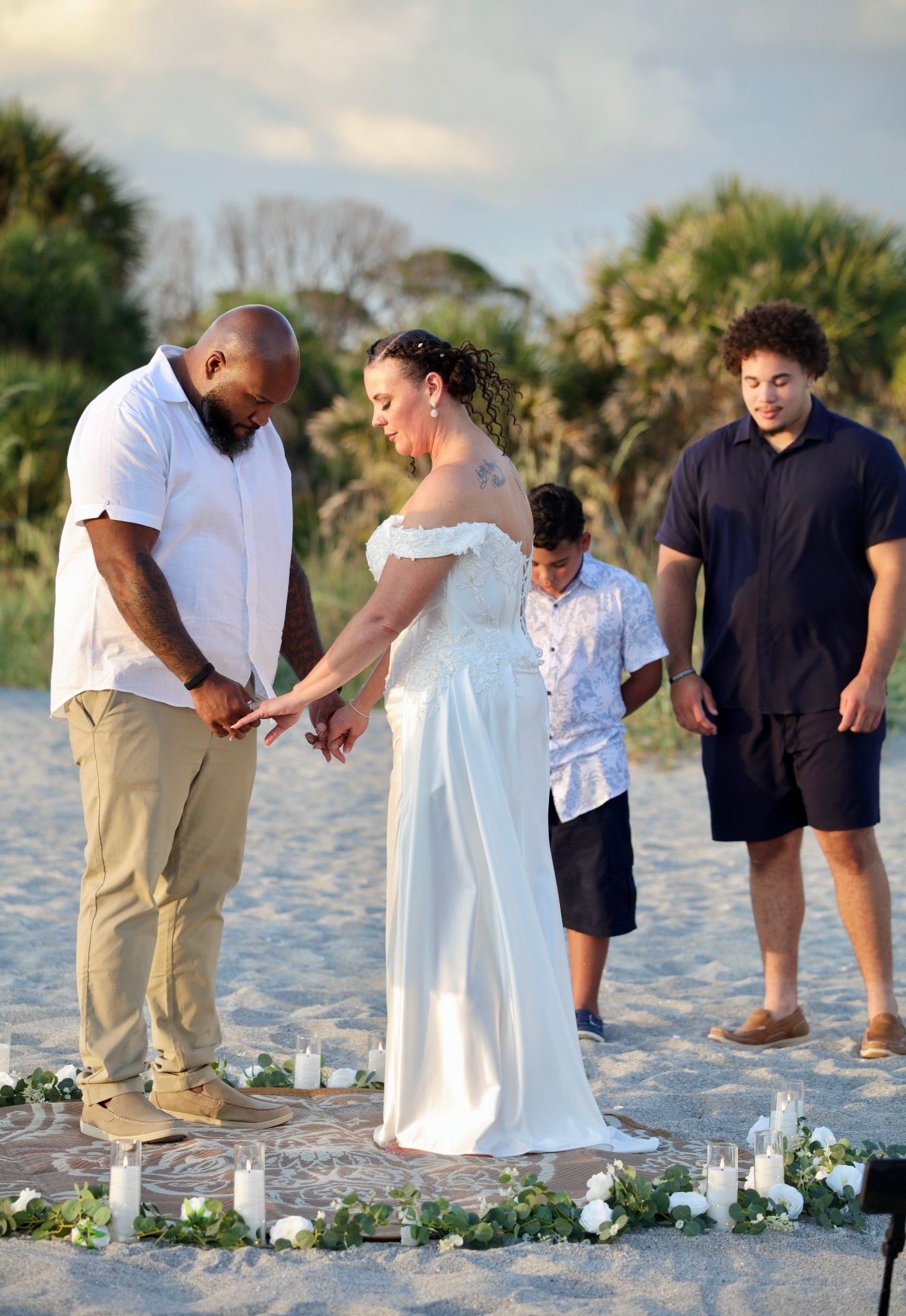 Beach wedding ceremony with couple exchanging rings, flanked by two children, candles and florals on sand.