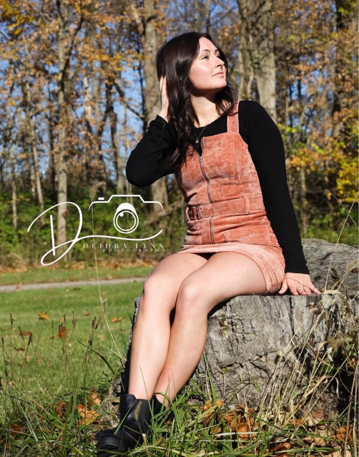 Young woman in rust overalls and black turtleneck sits on a rock outdoors in autumn park setting.