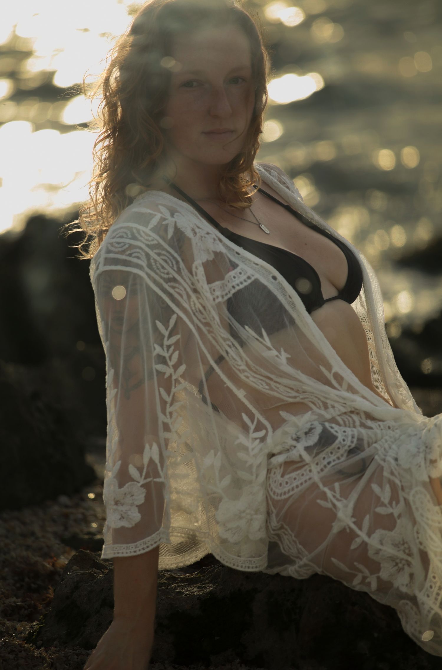 Woman in black bikini and white lace wrap sits on beach rocks with golden sunlight behind her.