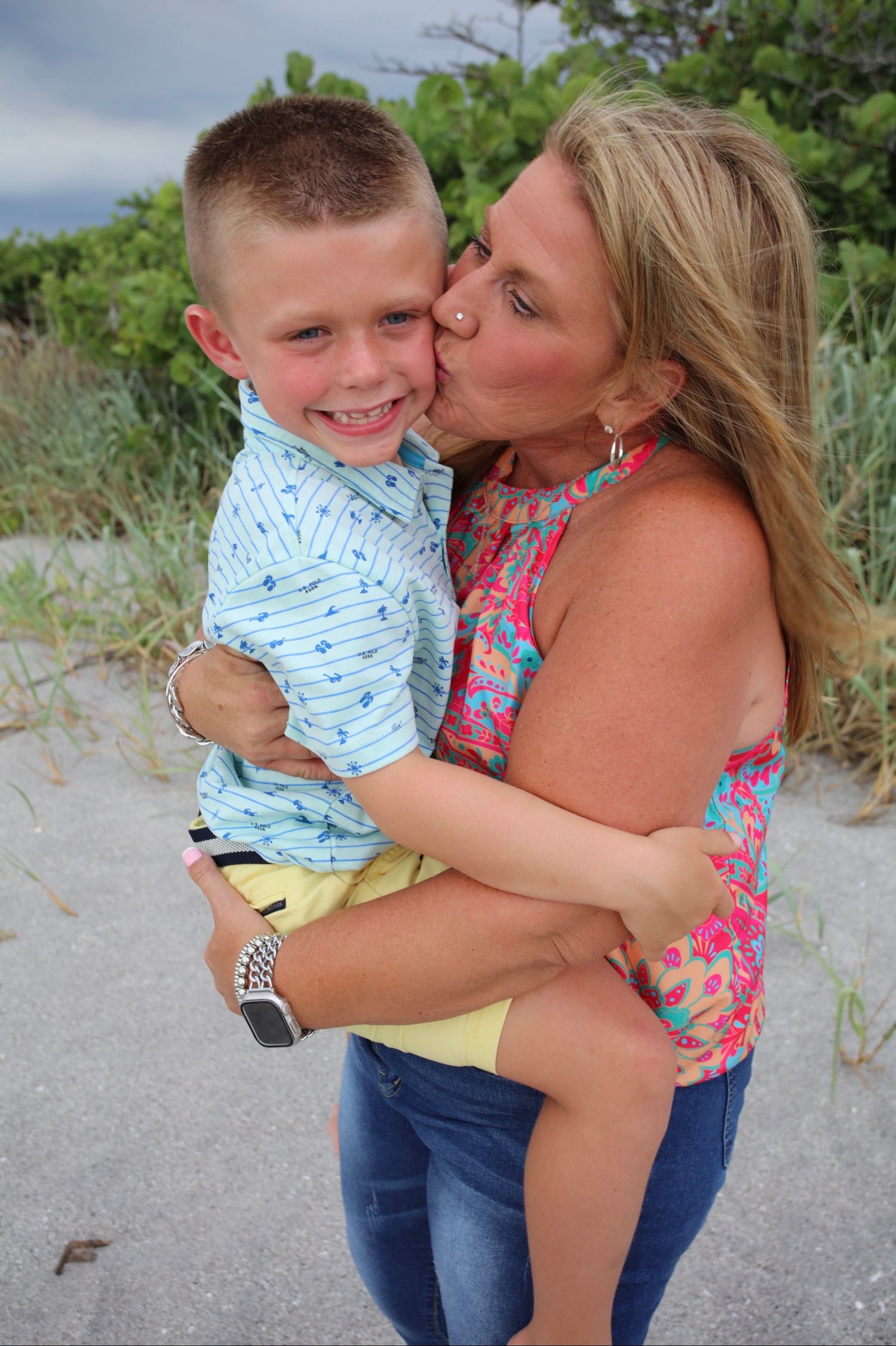 A woman in a floral top kisses a smiling boy in a blue shirt on a sandy beach.