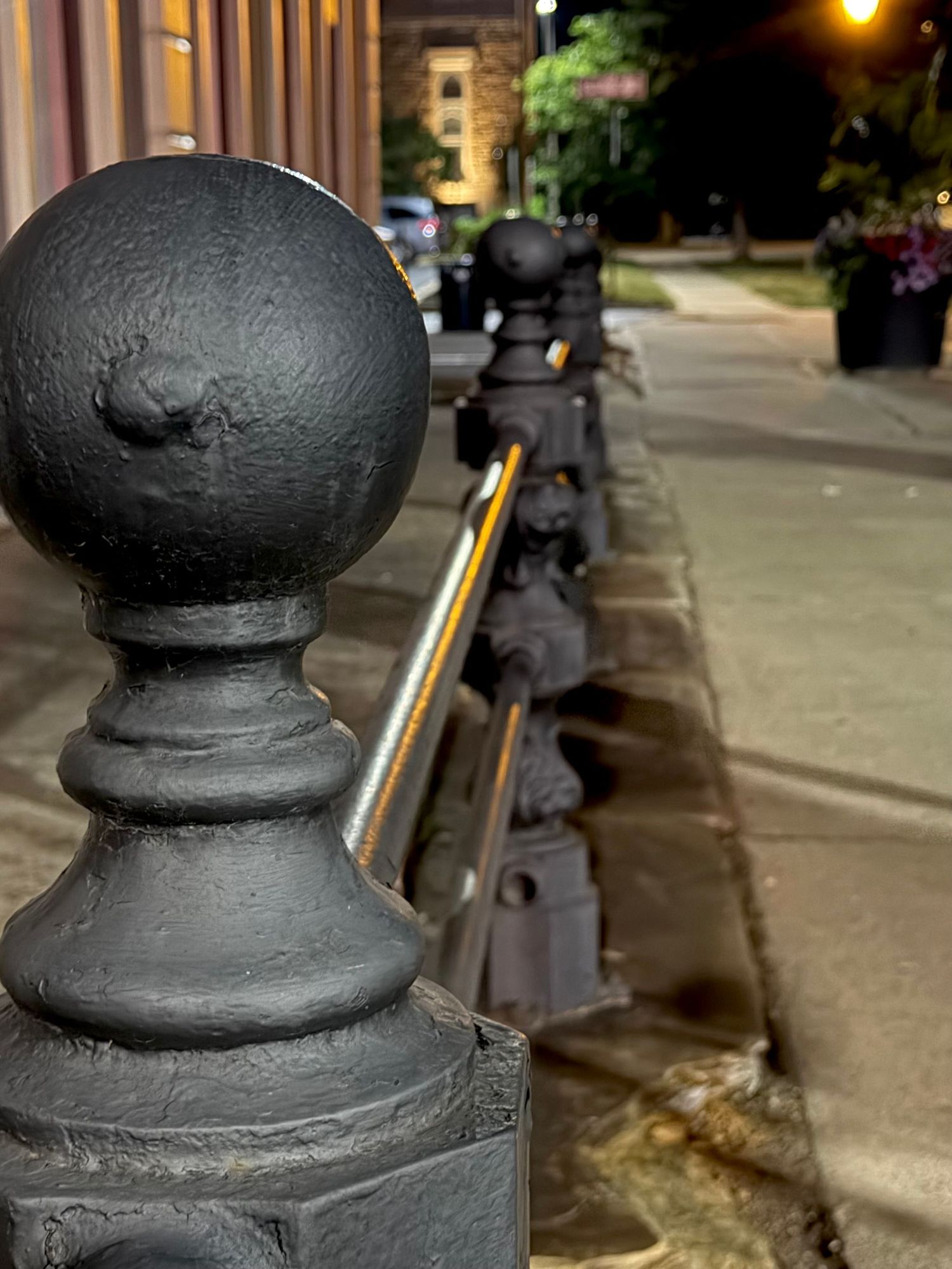 Black cast iron bollards with ornate ball tops line a city sidewalk at night, connected by a metal chain.