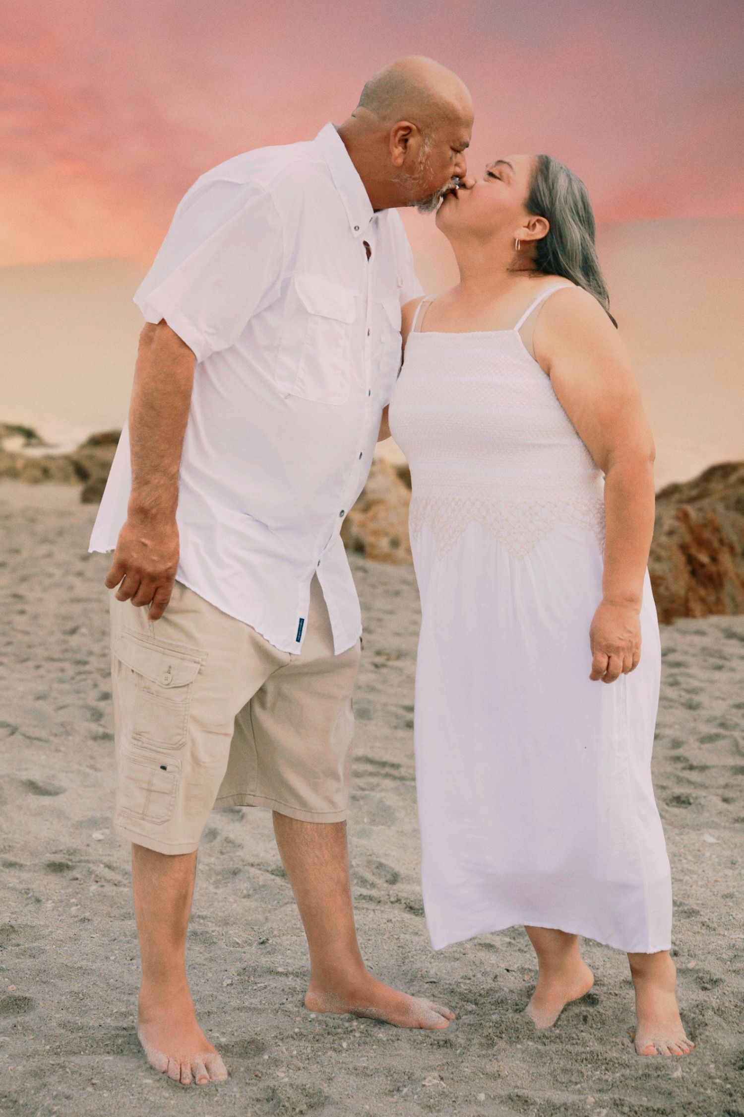 Couple kissing on beach at sunset, wearing white outfits, barefoot on sand with colorful sky.