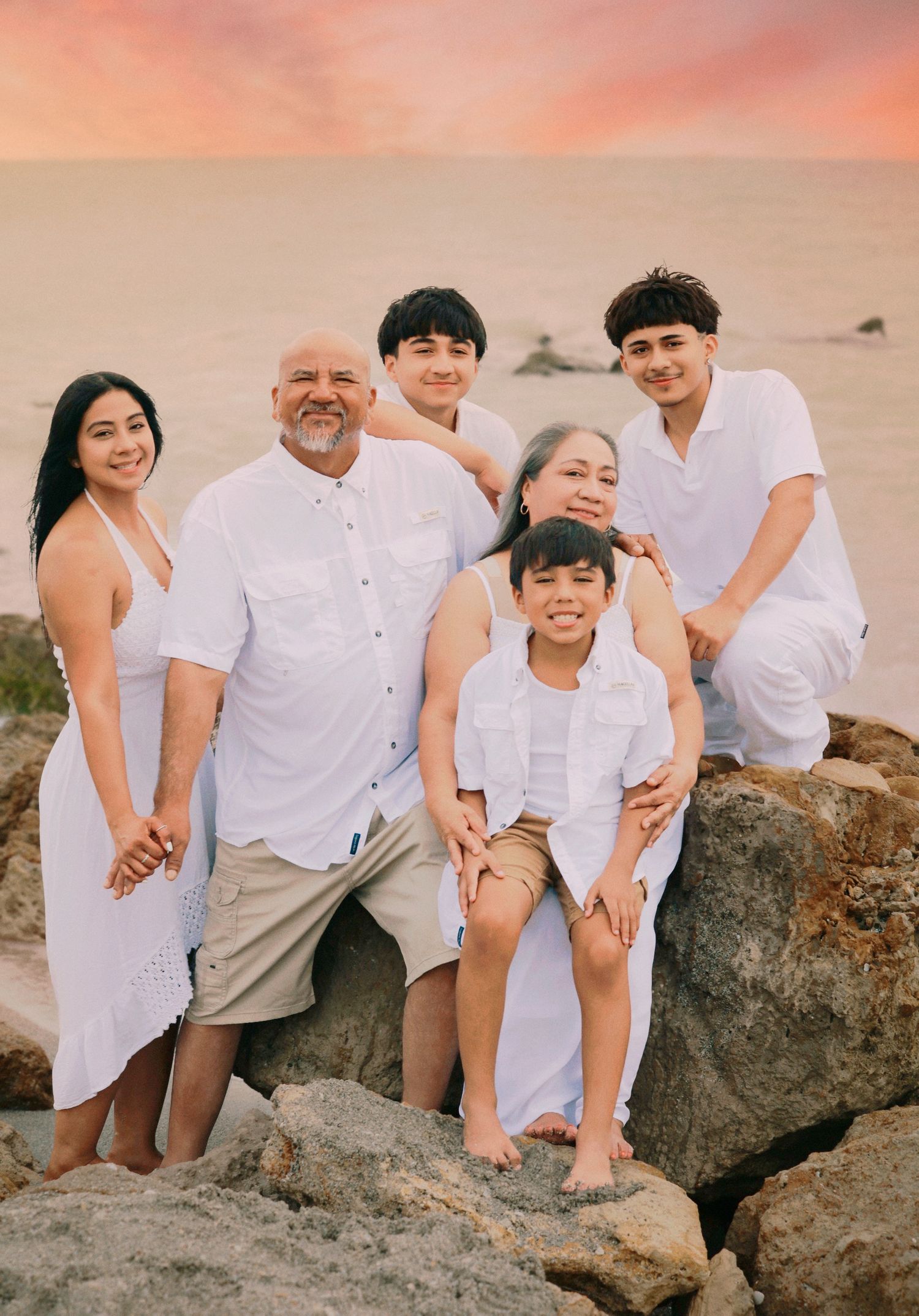 Family of six in white outfits posing on rocks at beach during sunset.
