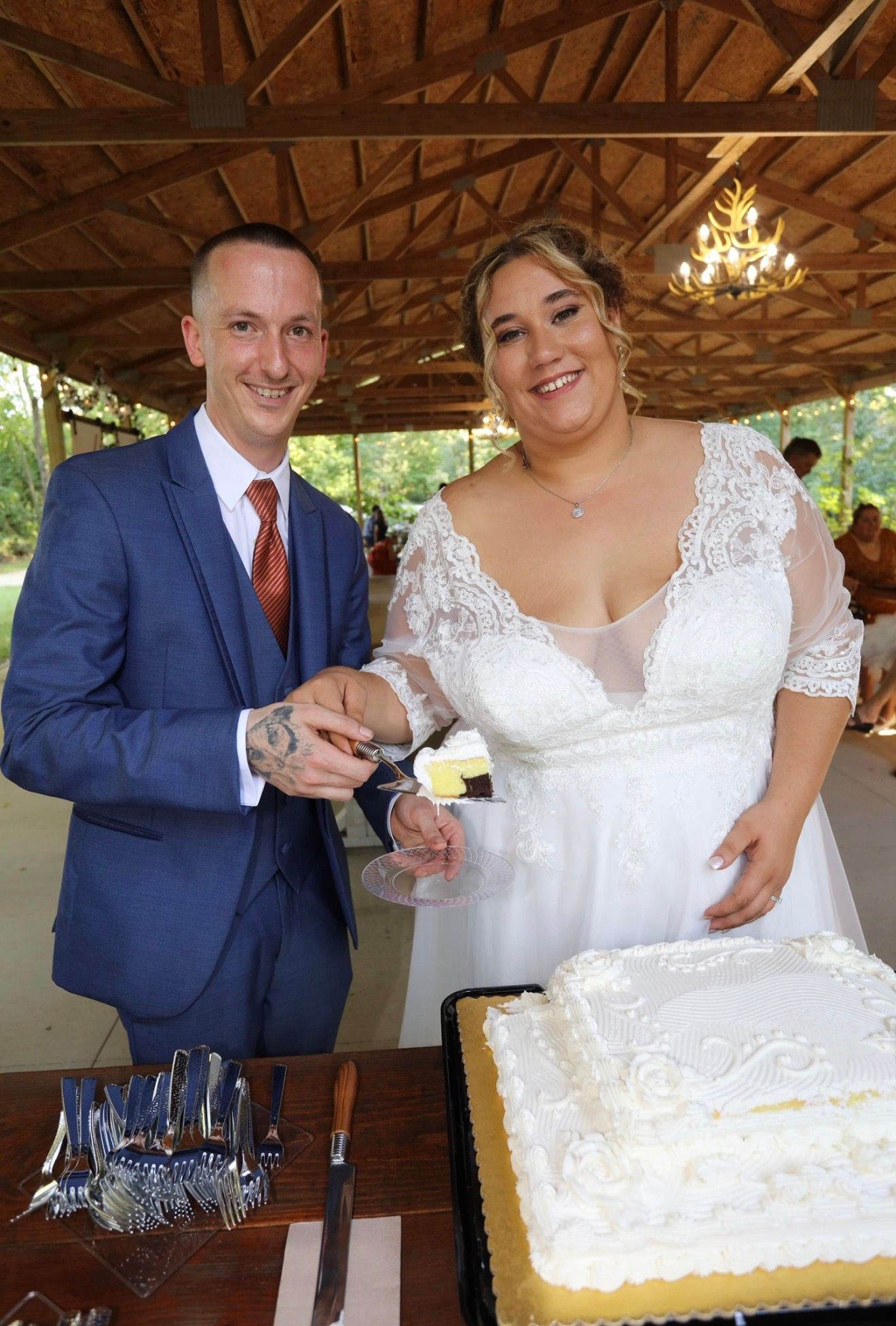 Bride and groom cutting wedding cake together at outdoor venue, bride in white lace gown, groom in blue suit.