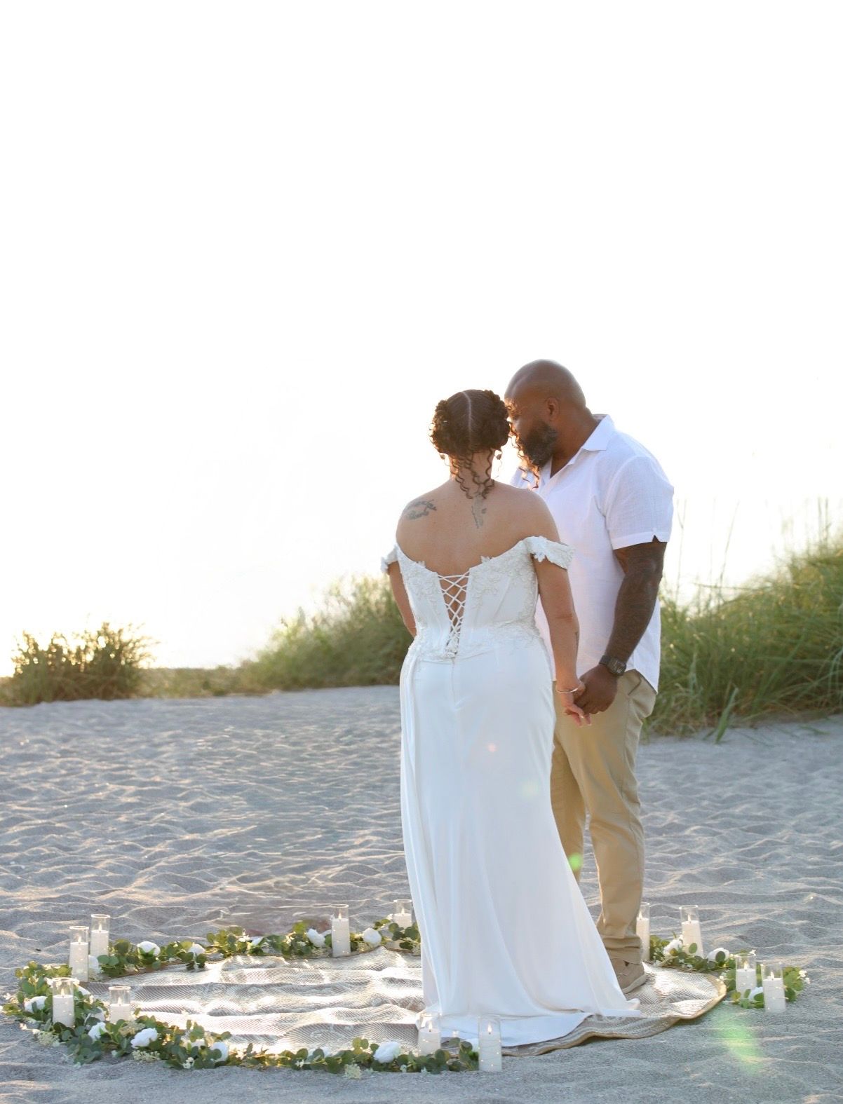 Newlyweds share intimate moment inside floral candle circle on beach at sunset.