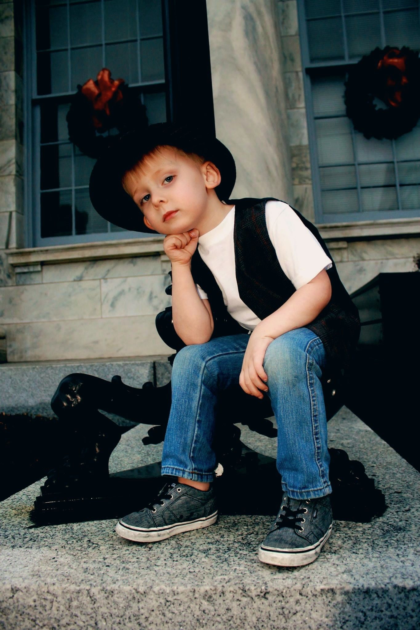 A young boy in a black fedora hat, vest, jeans, and sneakers sits thoughtfully on stone steps near Halloween decorations.