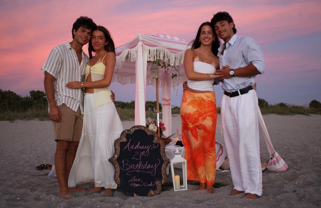 Two couples pose on a beach at sunset near a decorated canopy for a birthday celebration.