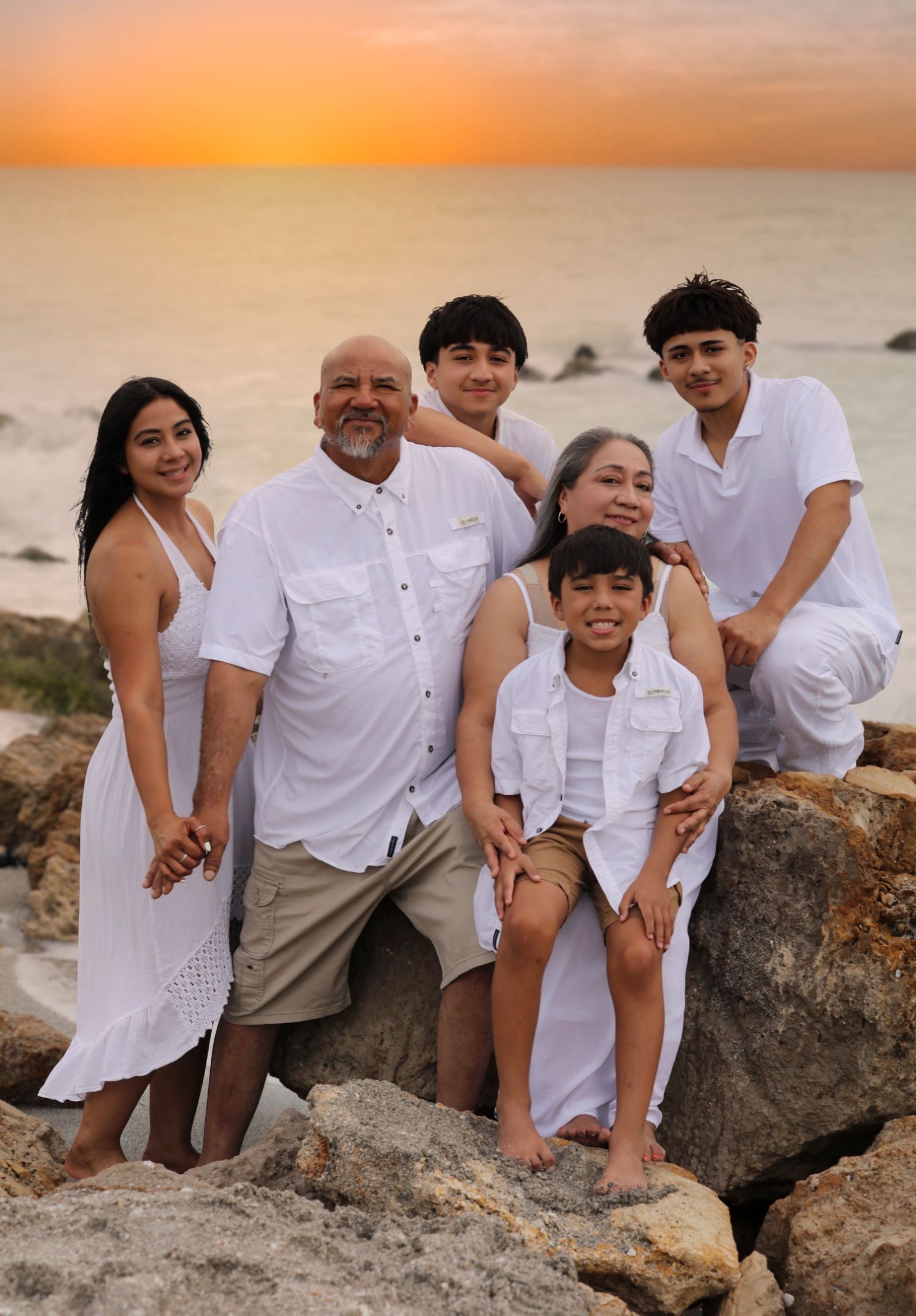 Family of six in white outfits posing on beach rocks at sunset with ocean backdrop.