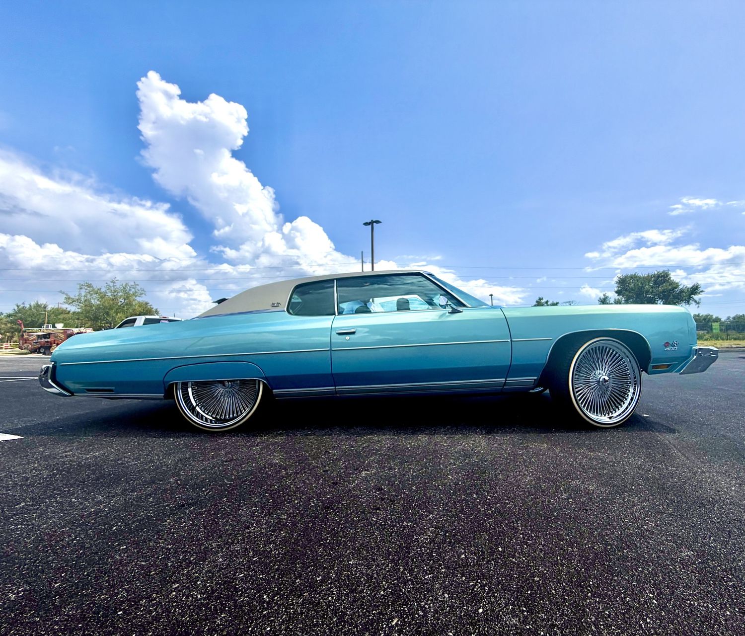 Vintage turquoise Cadillac lowrider with chrome wire wheels parked in a lot under a blue sky with white clouds.