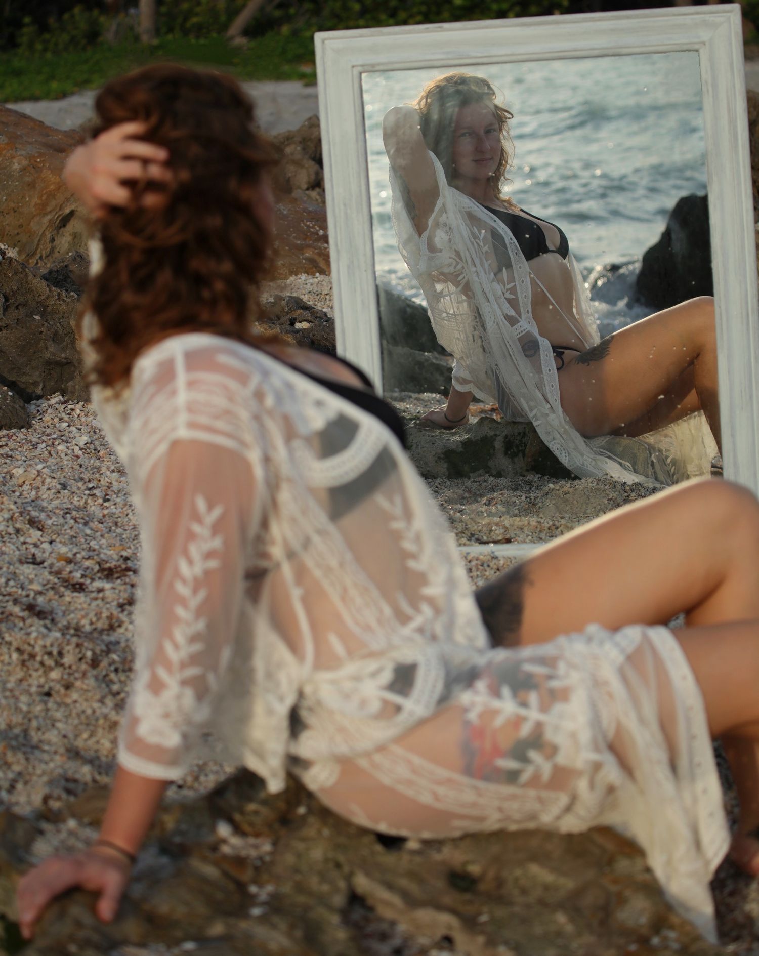Woman in sheer lace cover-up sitting on rocky beach looking at her reflection in a white-framed mirror.