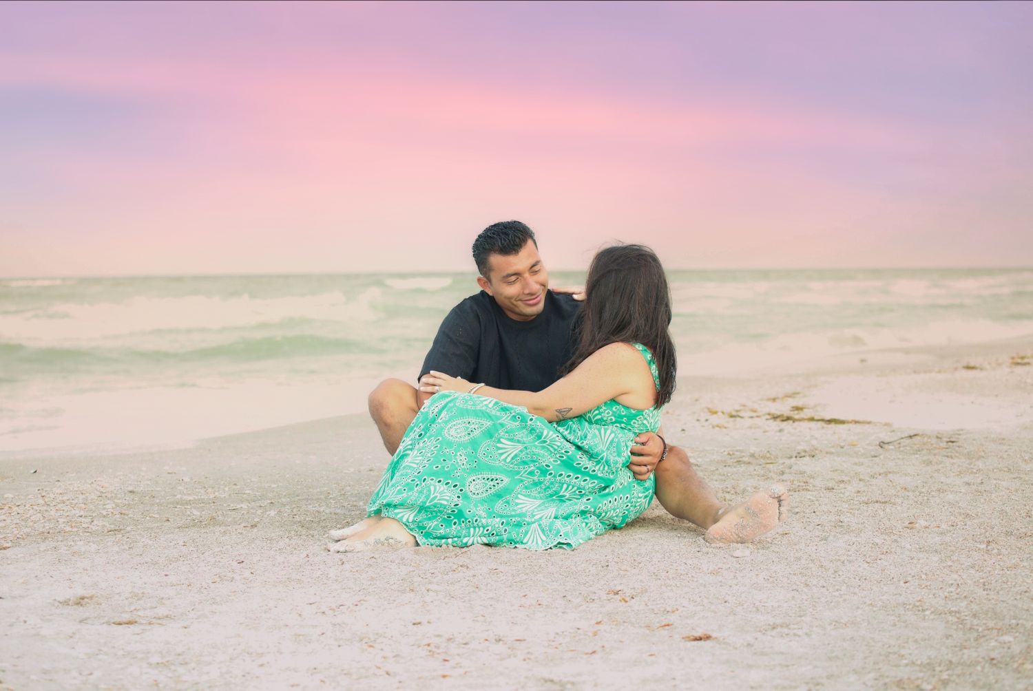 Couple sitting together on a sandy beach at sunset with a pink and purple sky and turquoise ocean waves.