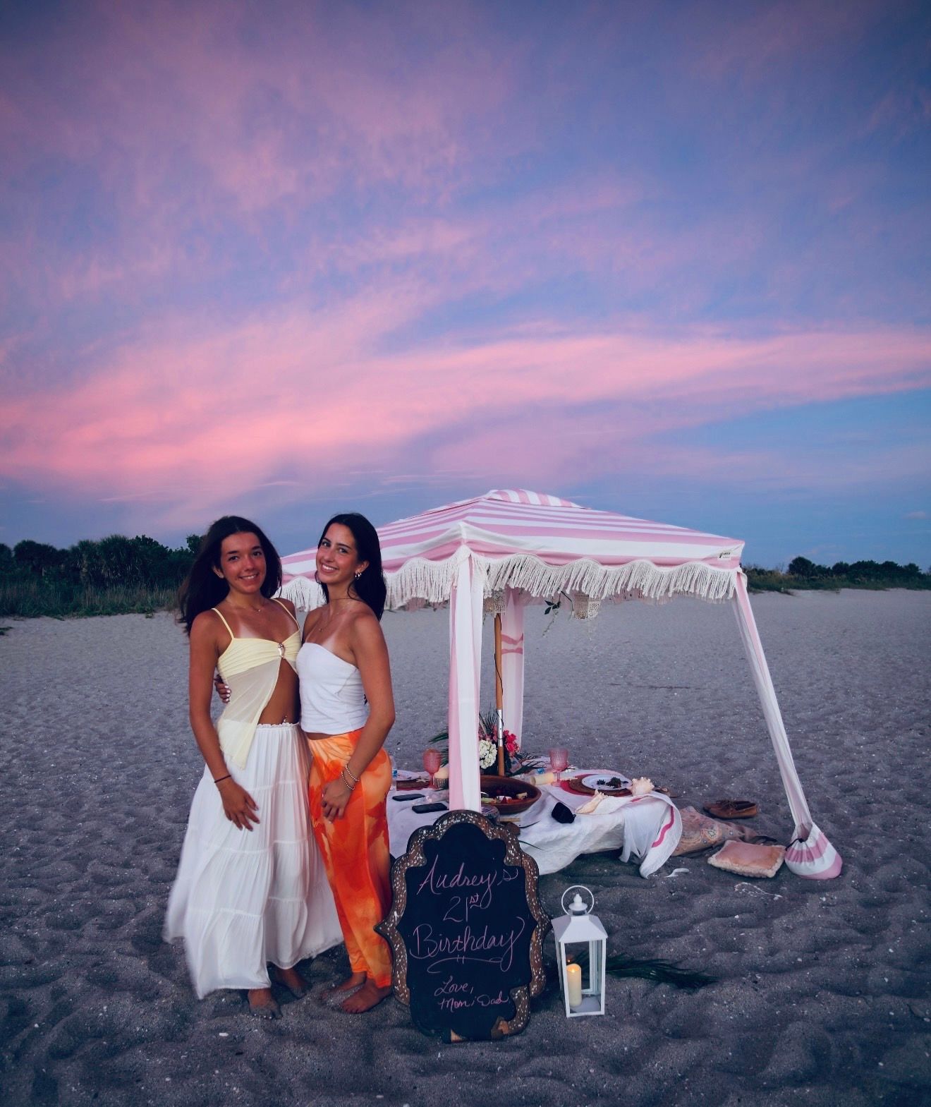 Two women pose at a beach birthday picnic setup under a pink fringe umbrella at sunset.