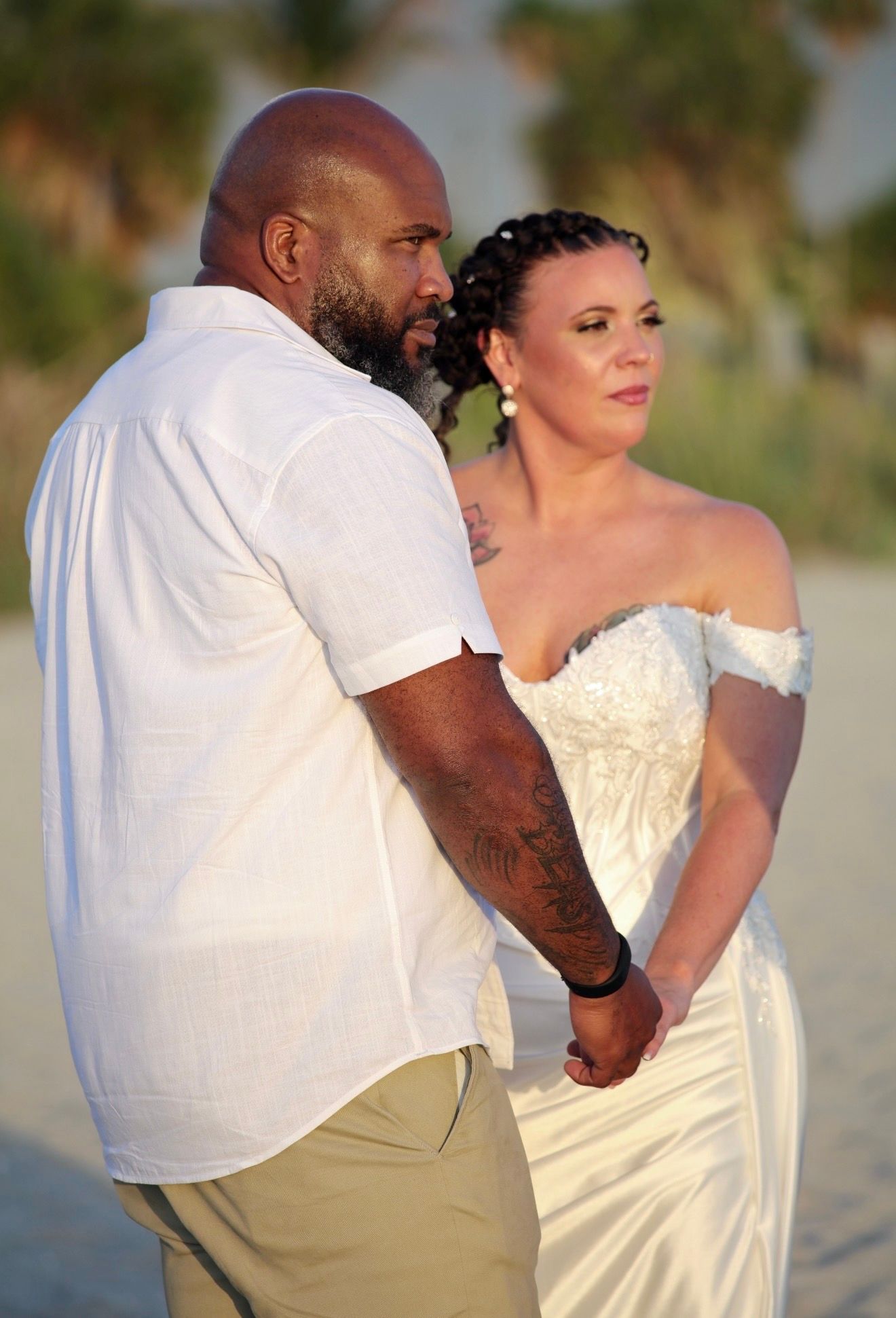Wedding couple holding hands on beach, bride in white off-shoulder gown, groom with tattoos in white shirt.