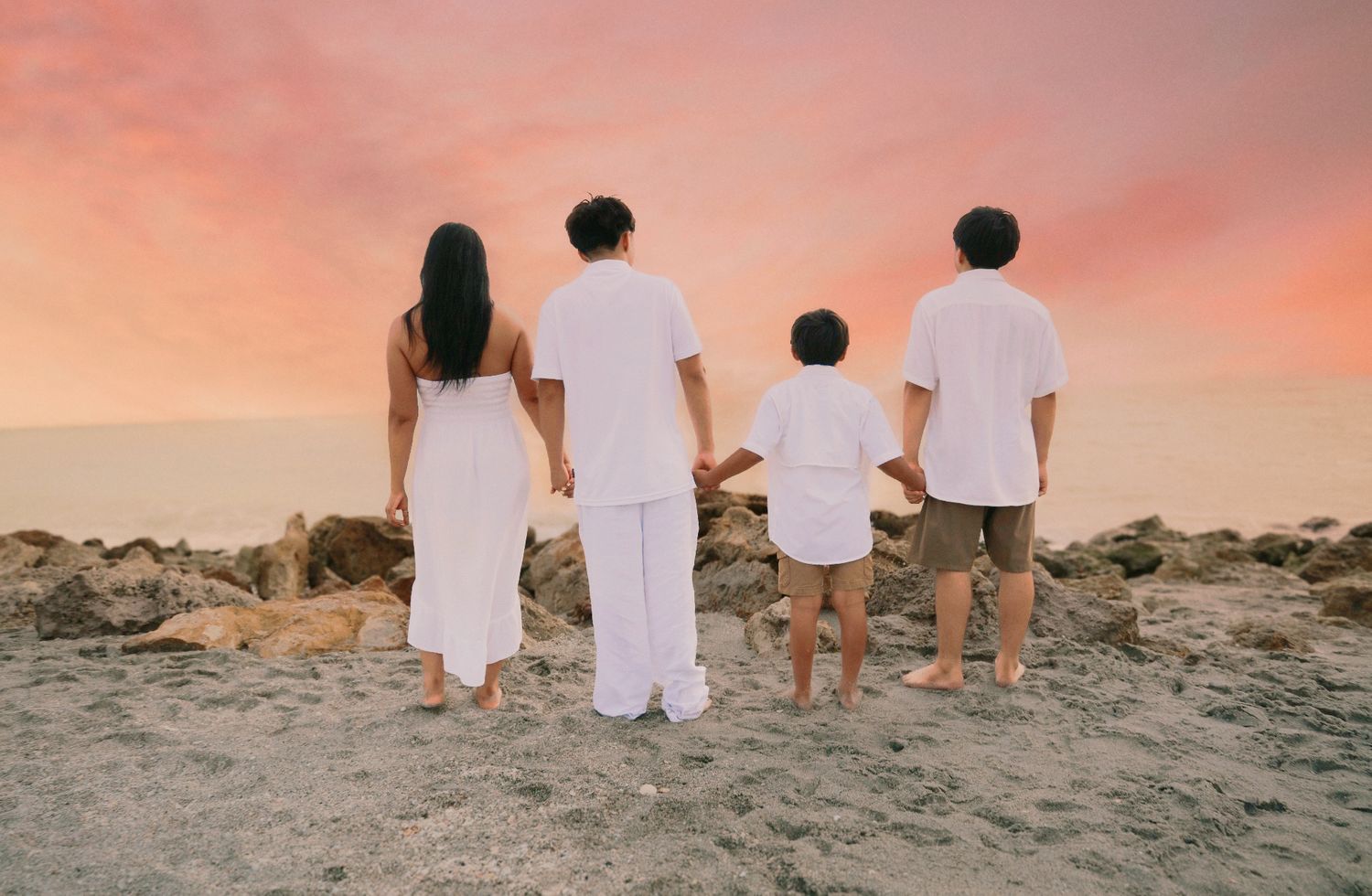 Family of four holding hands on rocky beach at sunset with pink and orange sky in background.