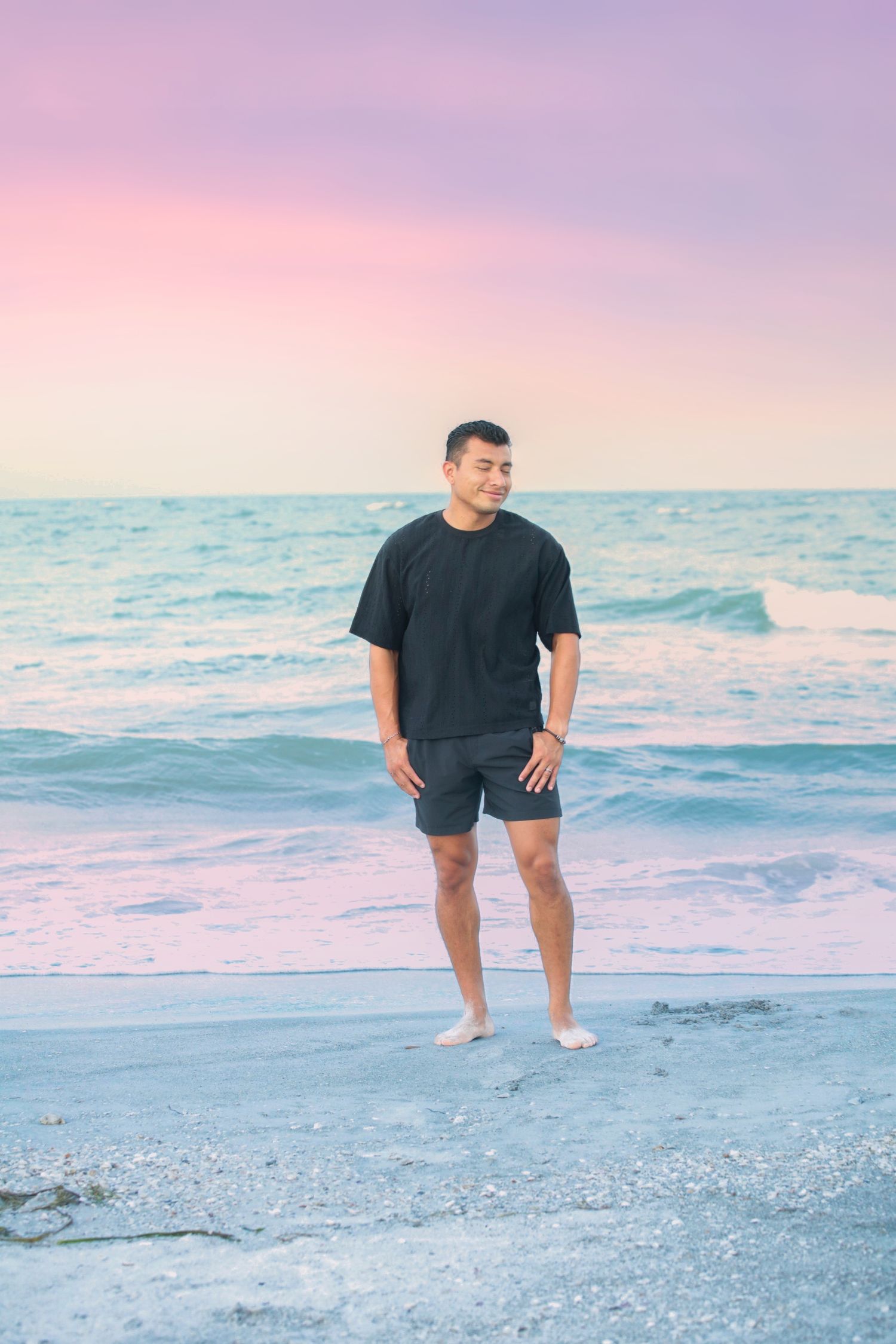 Man in black t-shirt and shorts standing barefoot on beach at sunset with pink and purple sky.