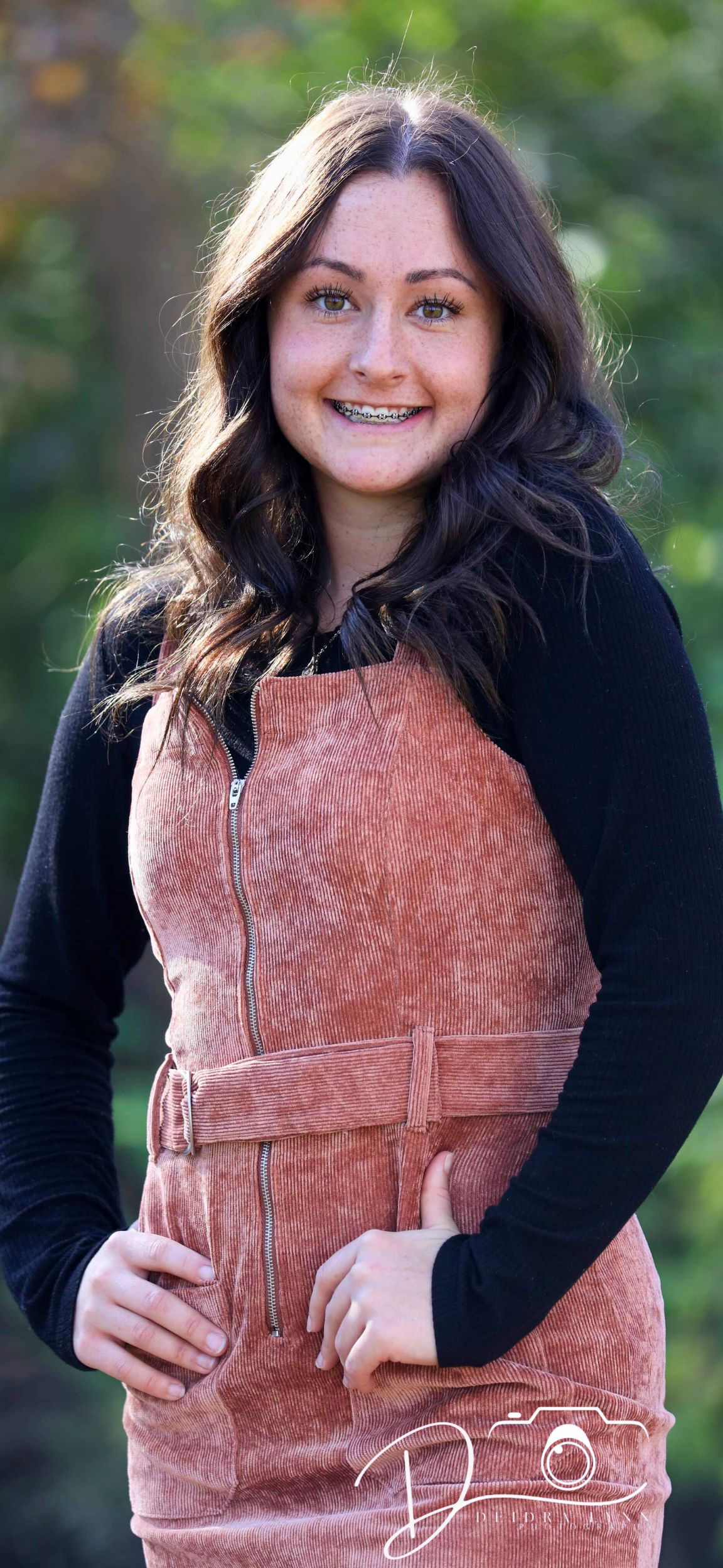 Young woman with dark hair and braces smiles outdoors, wearing a rust-colored zip-front dress over a black long-sleeve top.