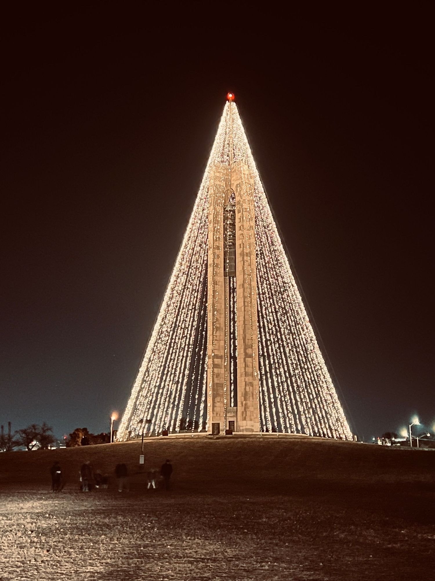 A towering Christmas tree made of lights illuminates the night sky at a festive outdoor monument display.