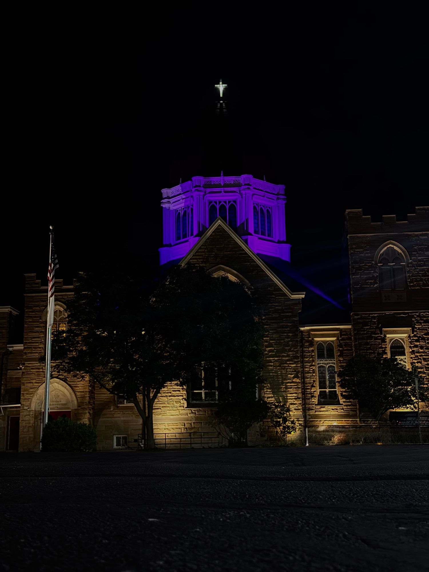 Historic stone church illuminated with purple lights at night, cross glowing atop the steeple against dark sky.