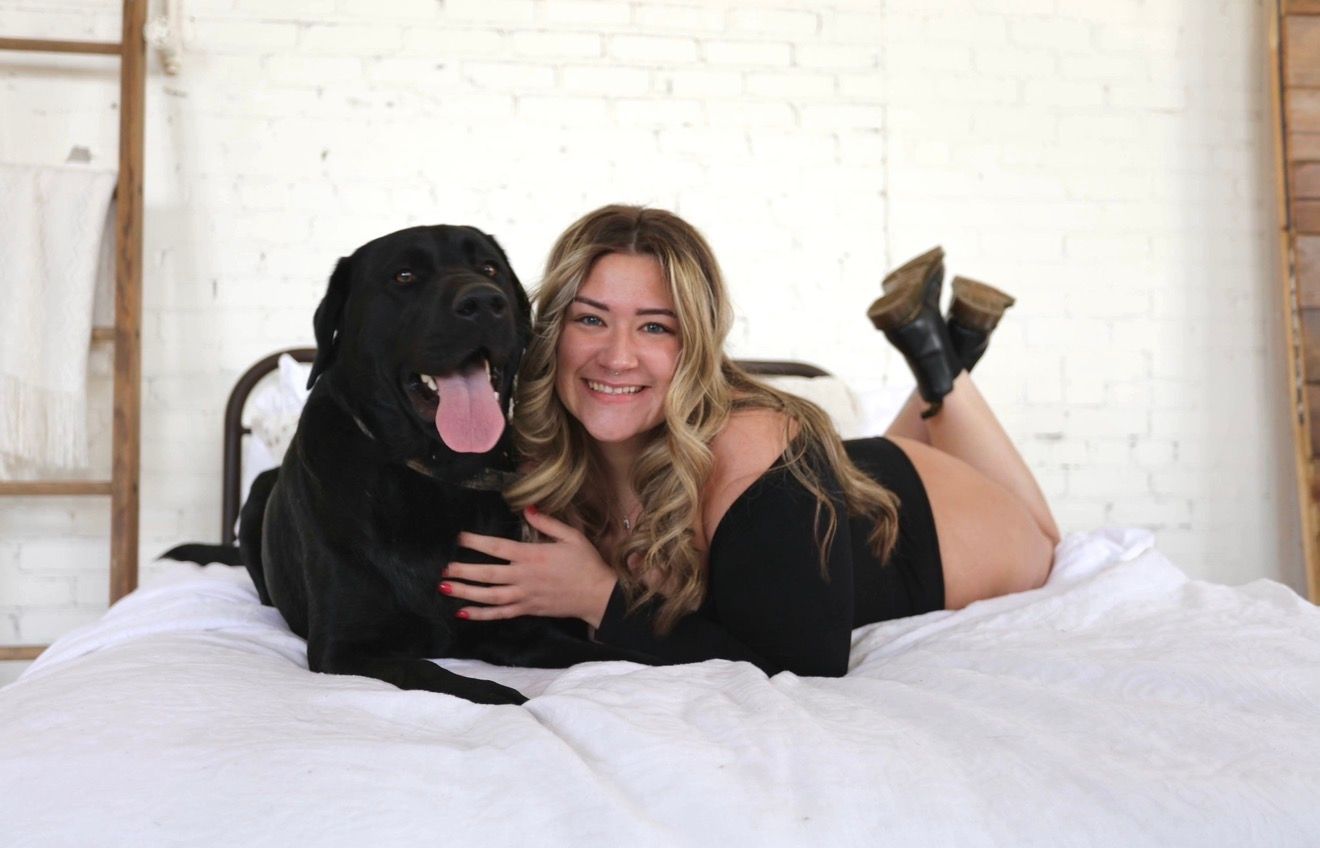 Woman smiling on white bed with black Labrador dog, both relaxed and happy in bright room with brick wall.