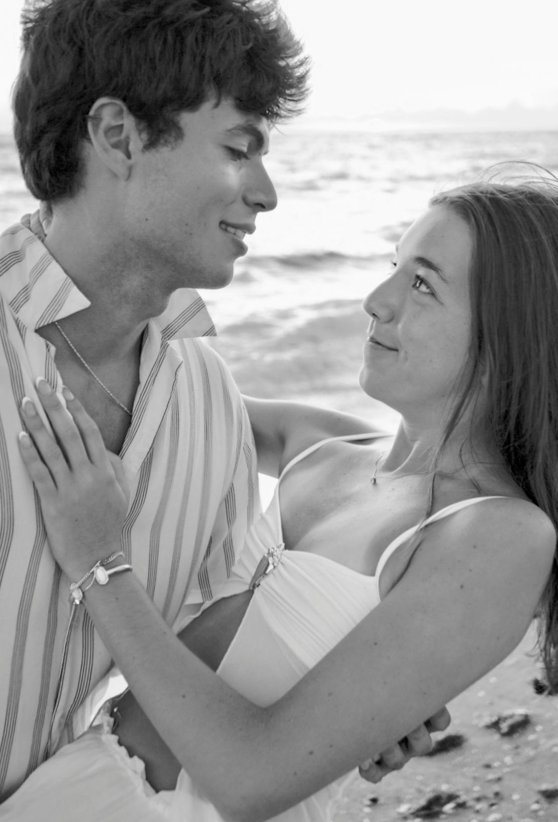 A romantic black and white photo of a young couple gazing at each other on the beach.