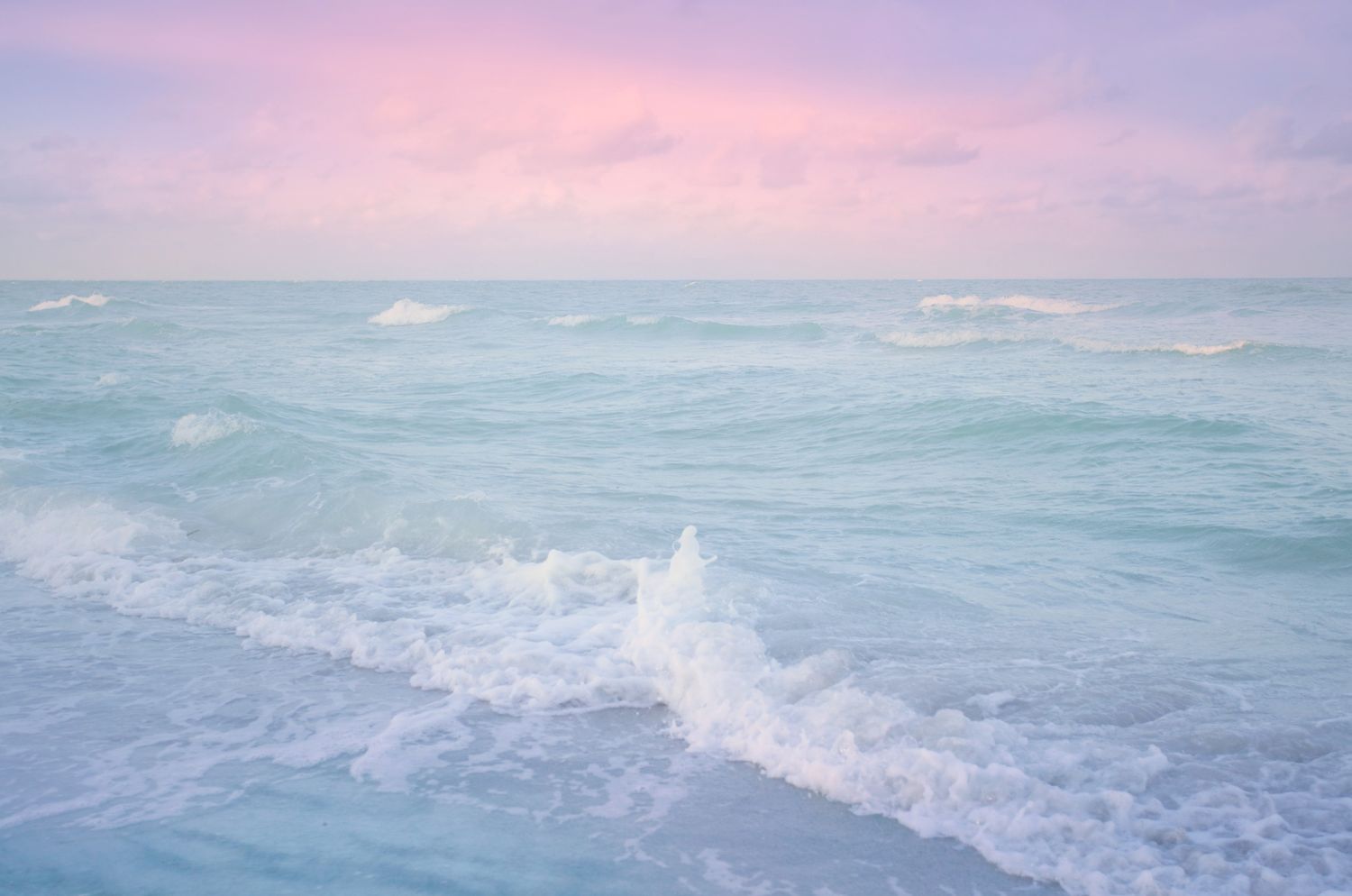 Pastel pink sky over turquoise ocean waves gently crashing on a serene beach at sunset.