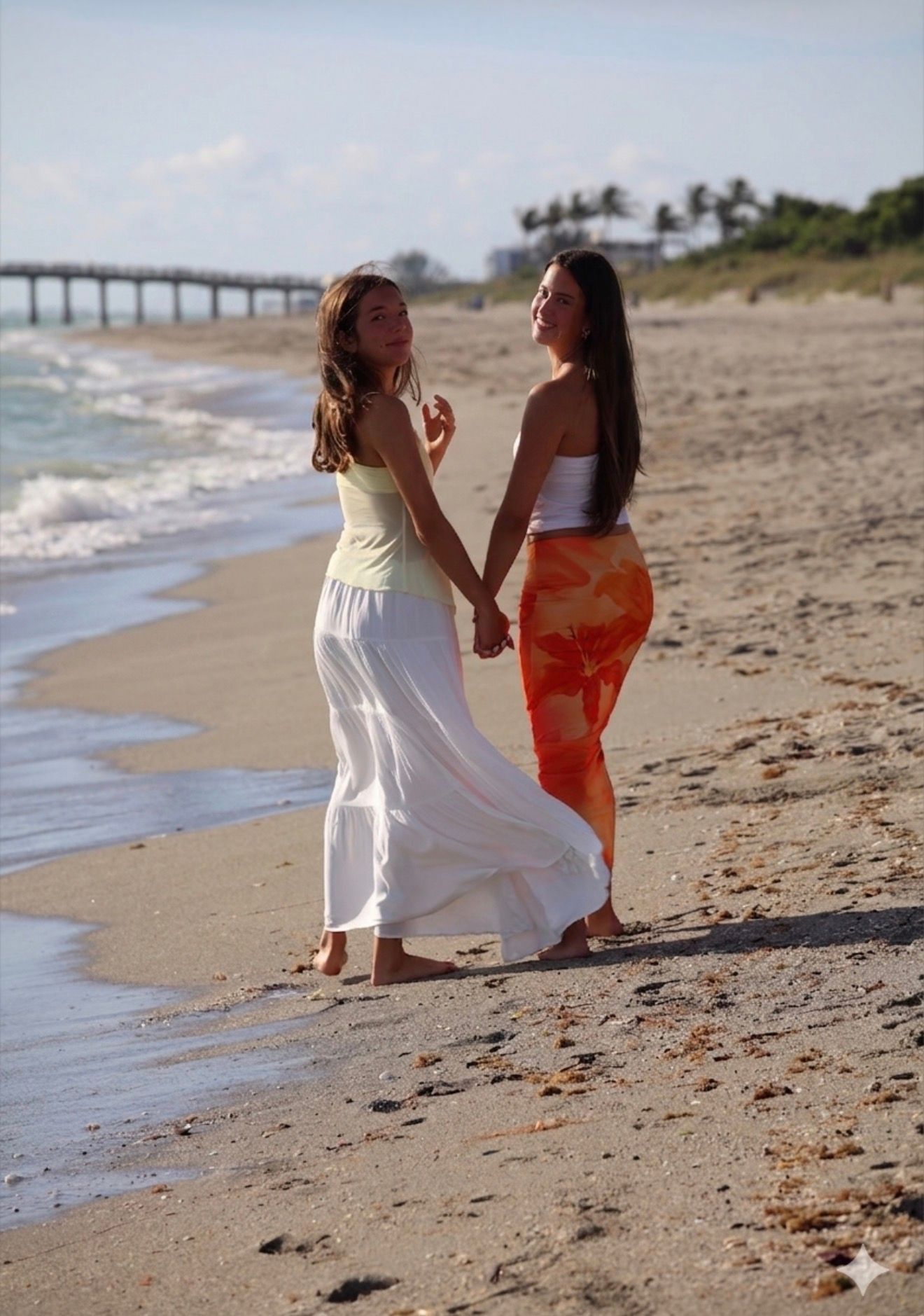 Two young women in flowing skirts hold hands while walking barefoot along a sunny beach near a pier.