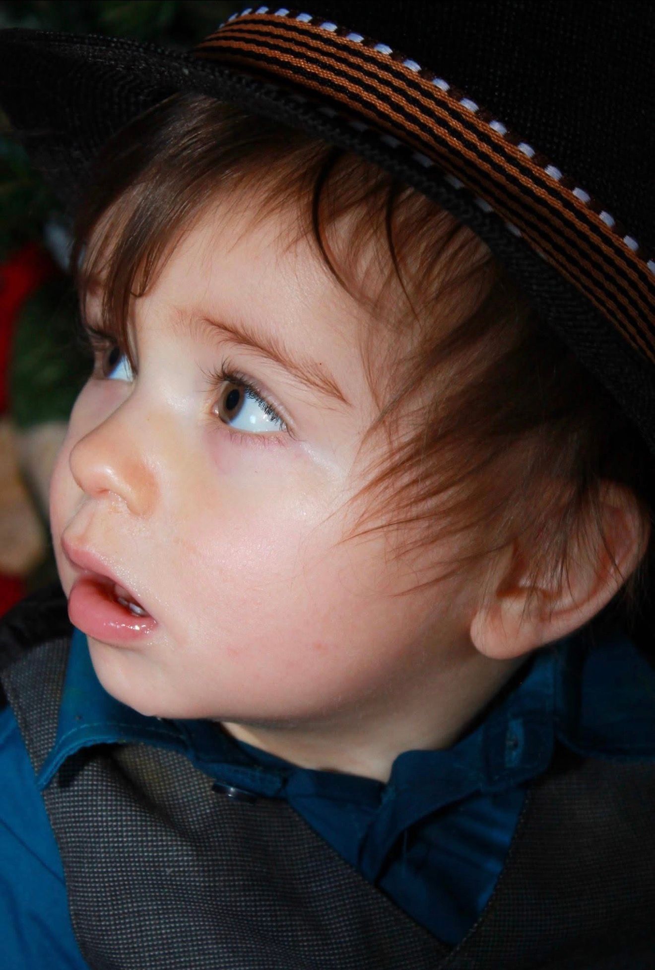 A young toddler wearing a black hat with orange stripes looks up curiously, dressed in a blue shirt.