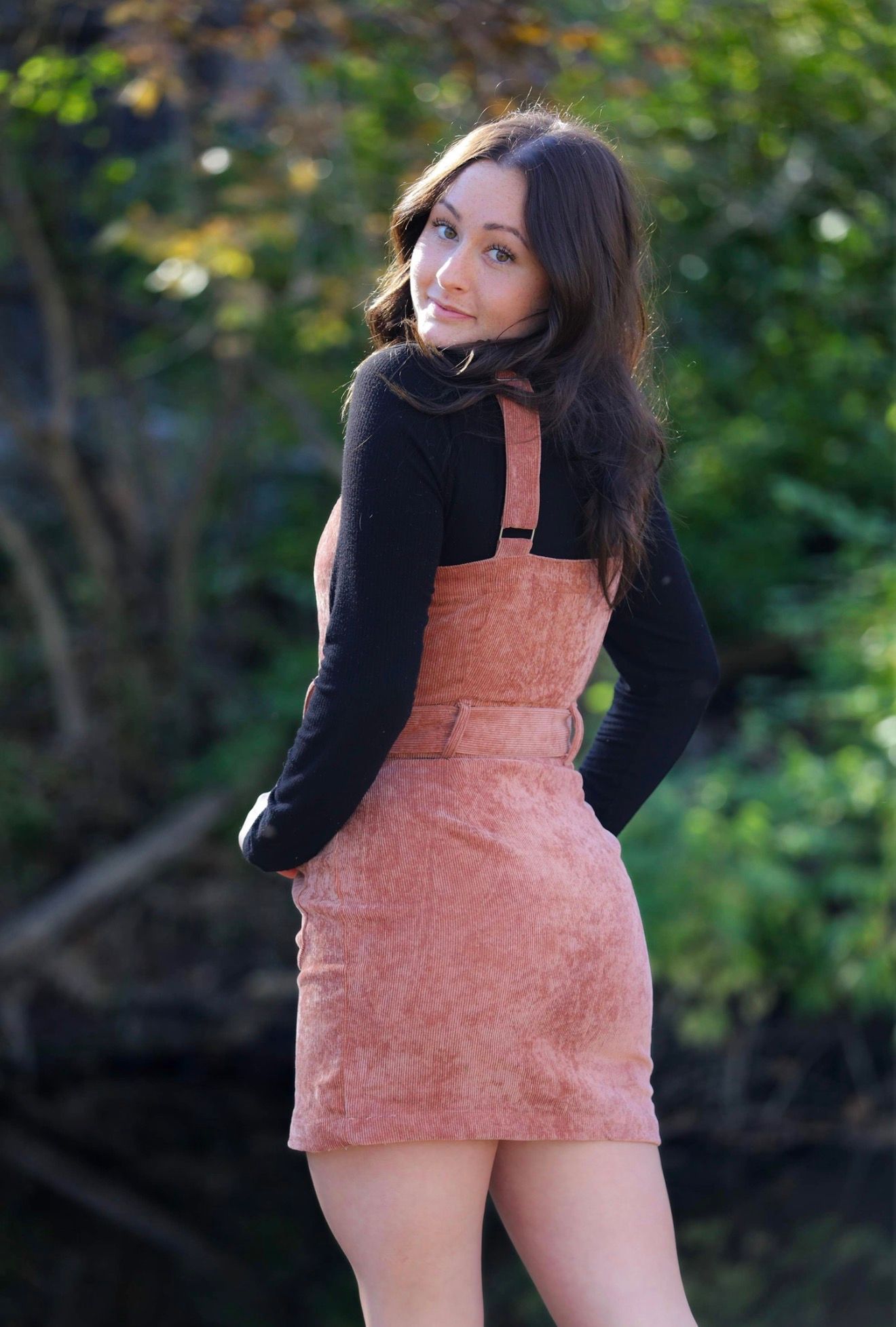 Young woman in pink corduroy pinafore dress and black top poses outdoors with green trees in background.