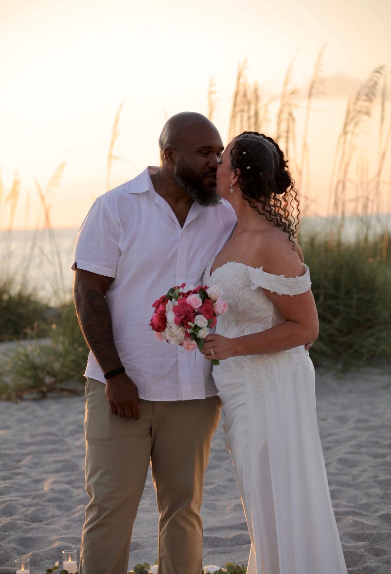 Newlyweds sharing a kiss on the beach at sunset, bride holding pink and white bouquet in white gown.