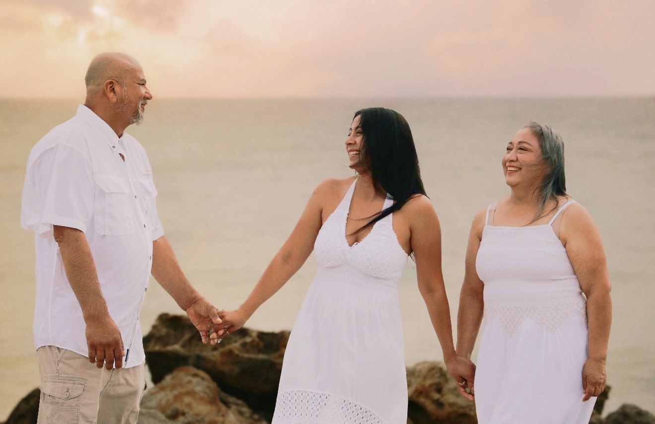 Three people in white attire hold hands on a rocky beach at sunset, smiling joyfully together.