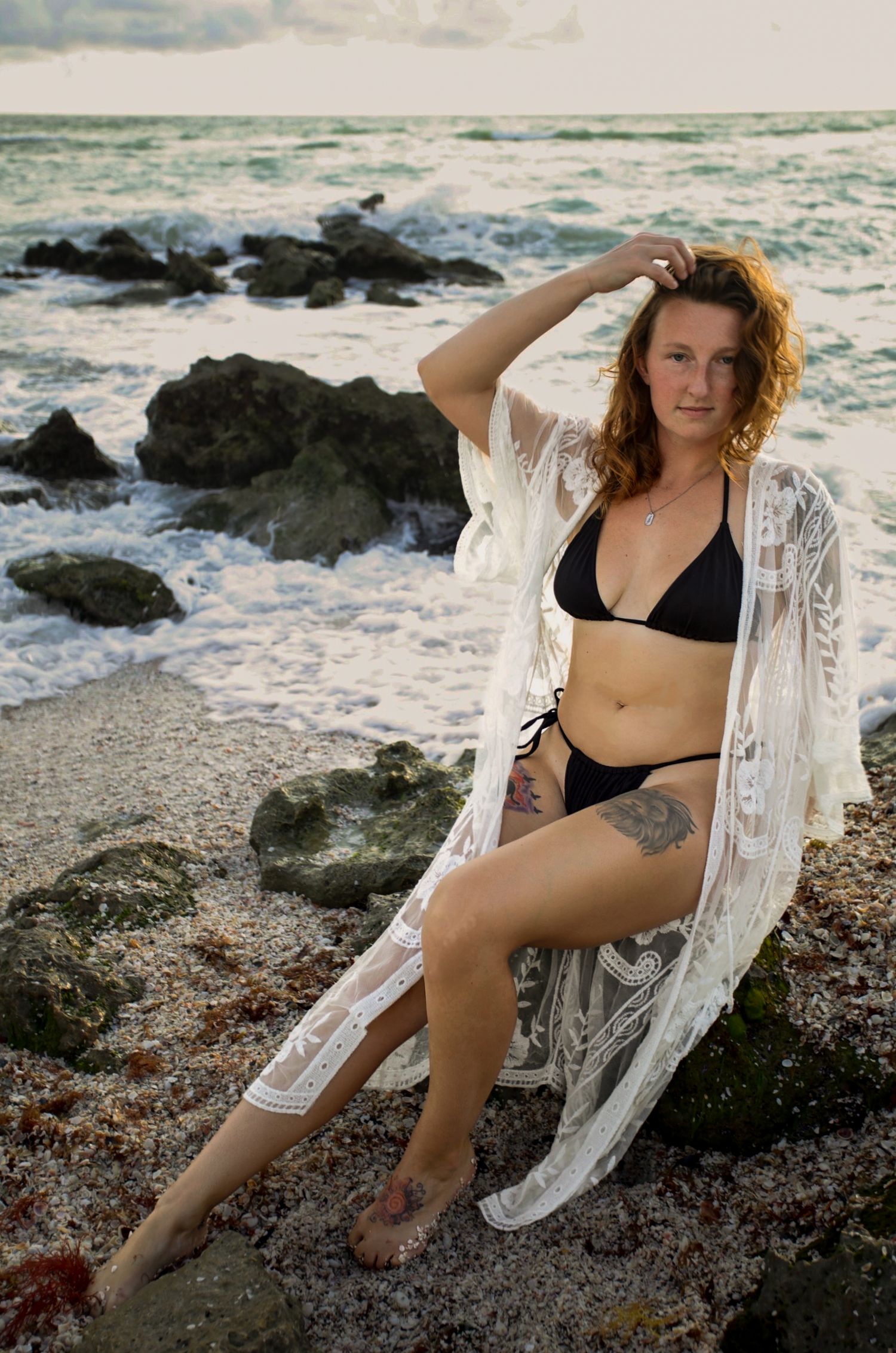 A woman in a black bikini and white lace cover-up sits on rocky shore with ocean waves behind her.