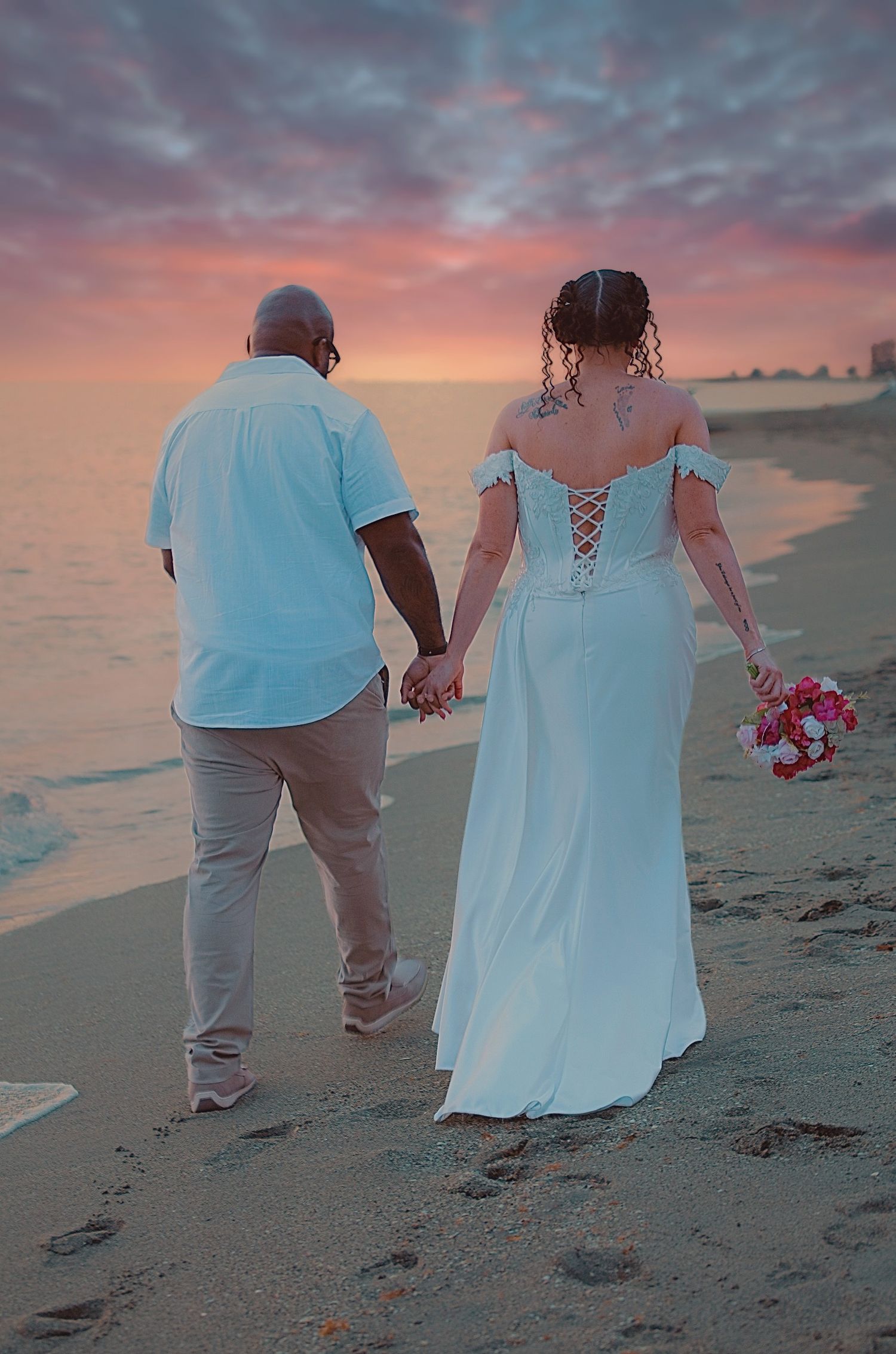 Newlywed couple holding hands walking on beach at sunset, bride in white lace gown holding bouquet.