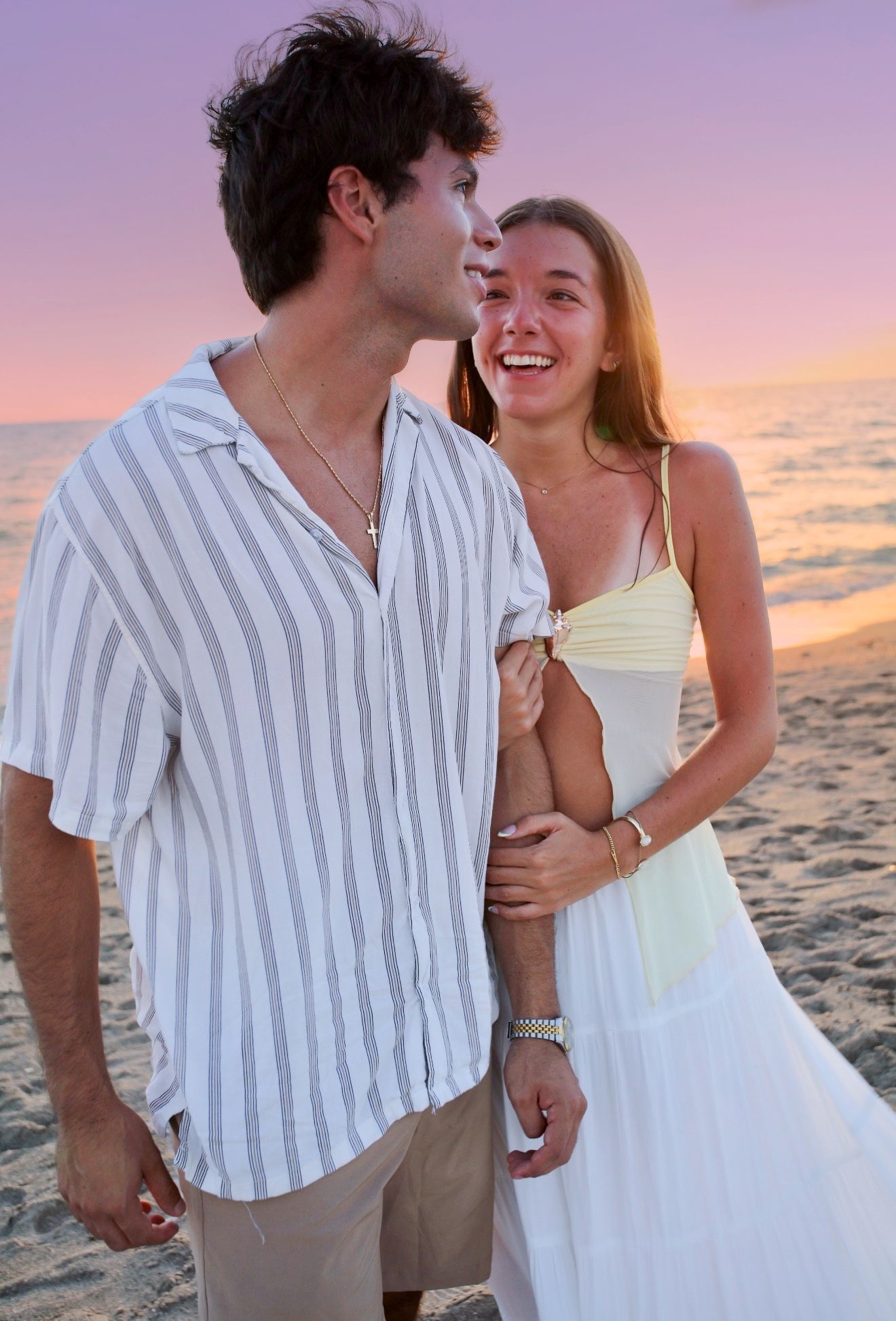 Happy young couple laughing together on sandy beach during vibrant pink and purple sunset.