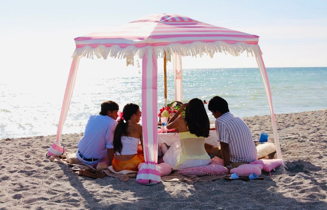 Family of four enjoying a picnic under a pink striped canopy on a sunny beach by the ocean.