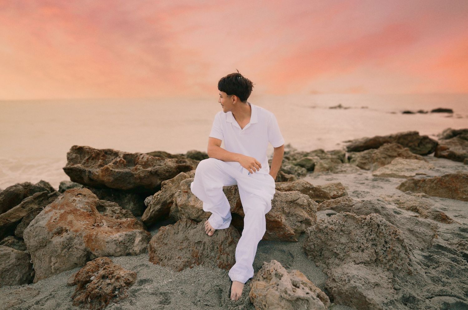 A young man in white outfit sits on rocky shore during a stunning pink and orange sunset over the ocean.