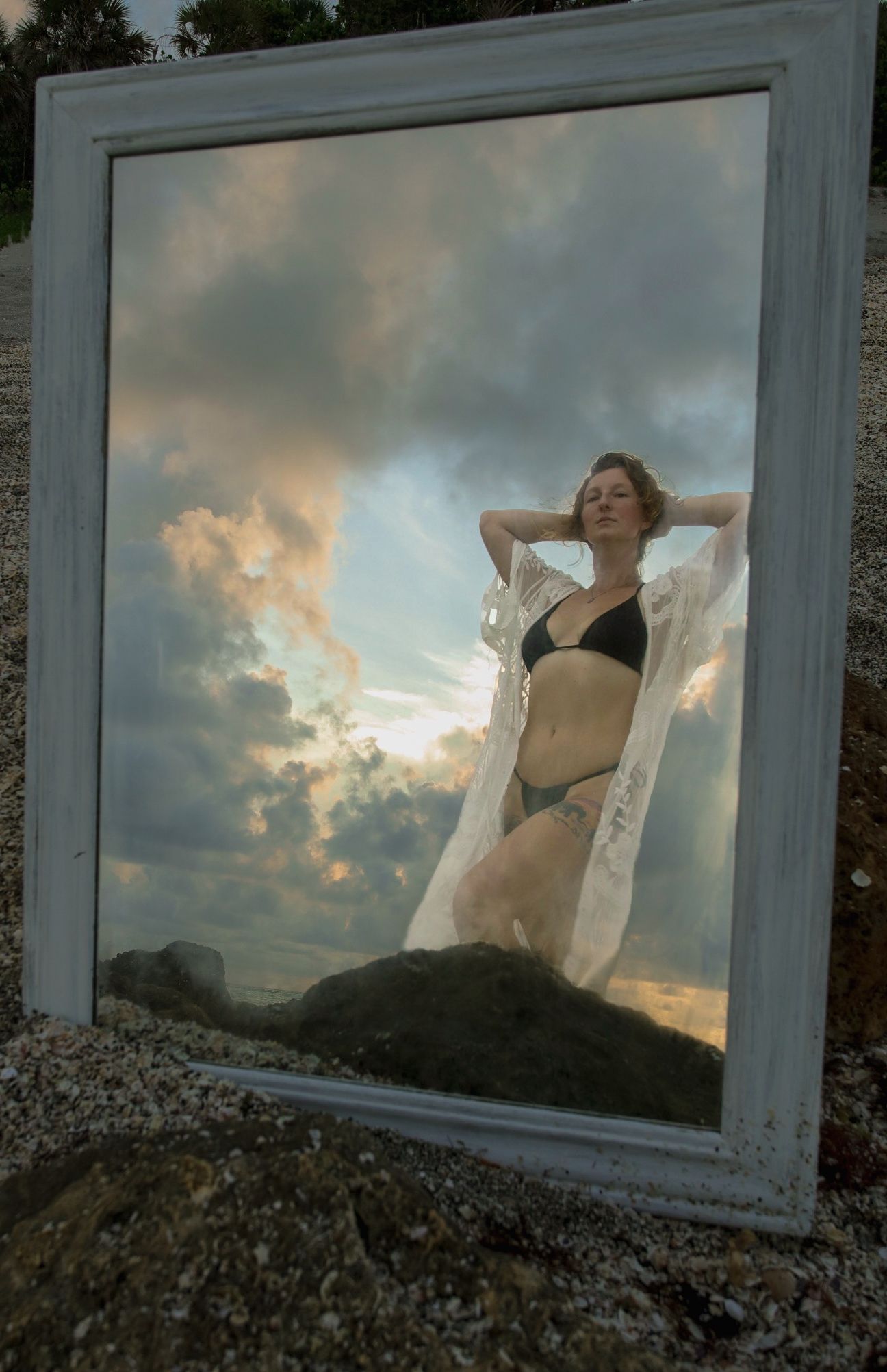 Woman in black bikini poses dramatically against cloudy sunset sky, framed in a rustic white picture frame on rocky beach.