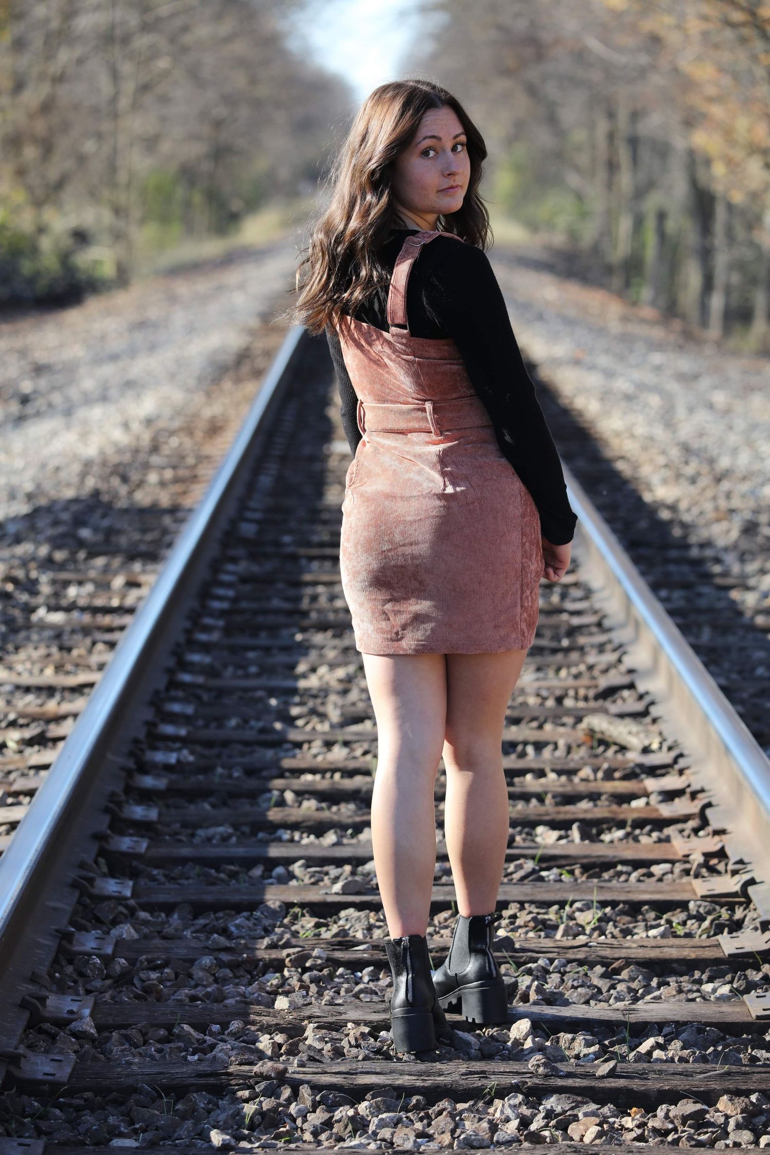 Young woman in pink dress and black top stands on railroad tracks, looking back over her shoulder on a sunny day.