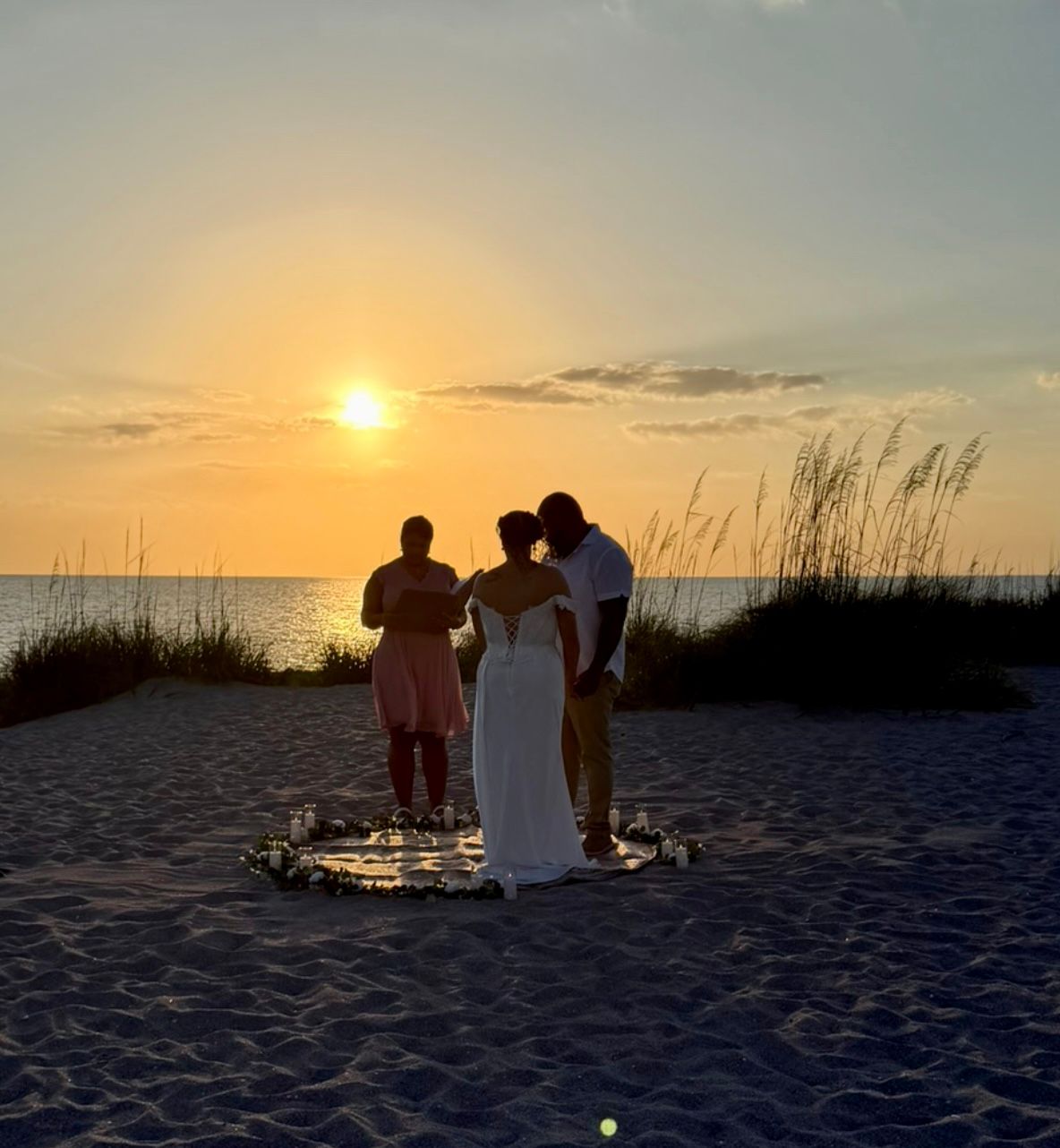 Couple exchanging vows at a stunning beach sunset wedding ceremony with sea oats and a floral circle arrangement.