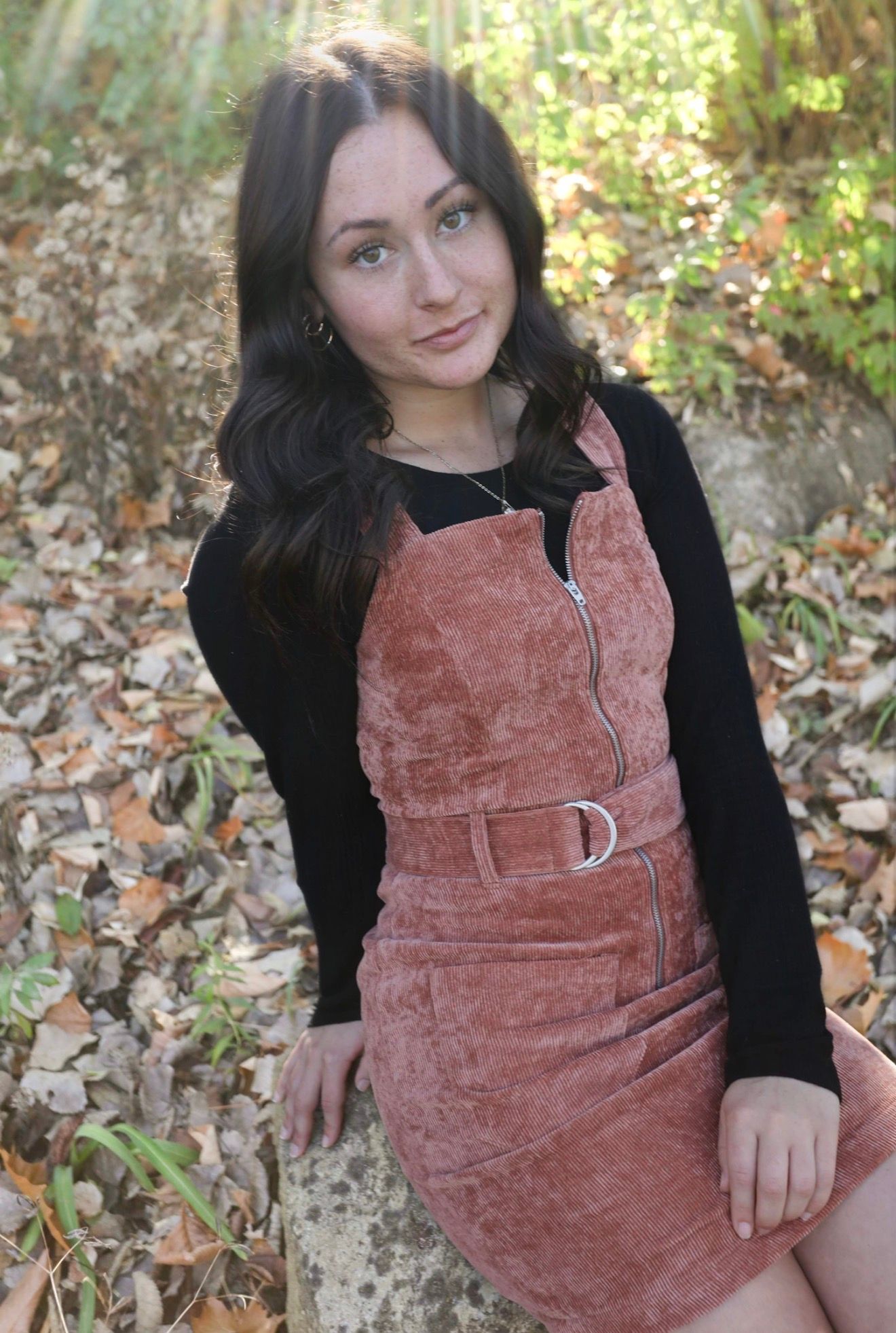 Young woman with dark hair wearing a pink velvet pinafore dress over black long-sleeve top, posing outdoors among autumn leaves.